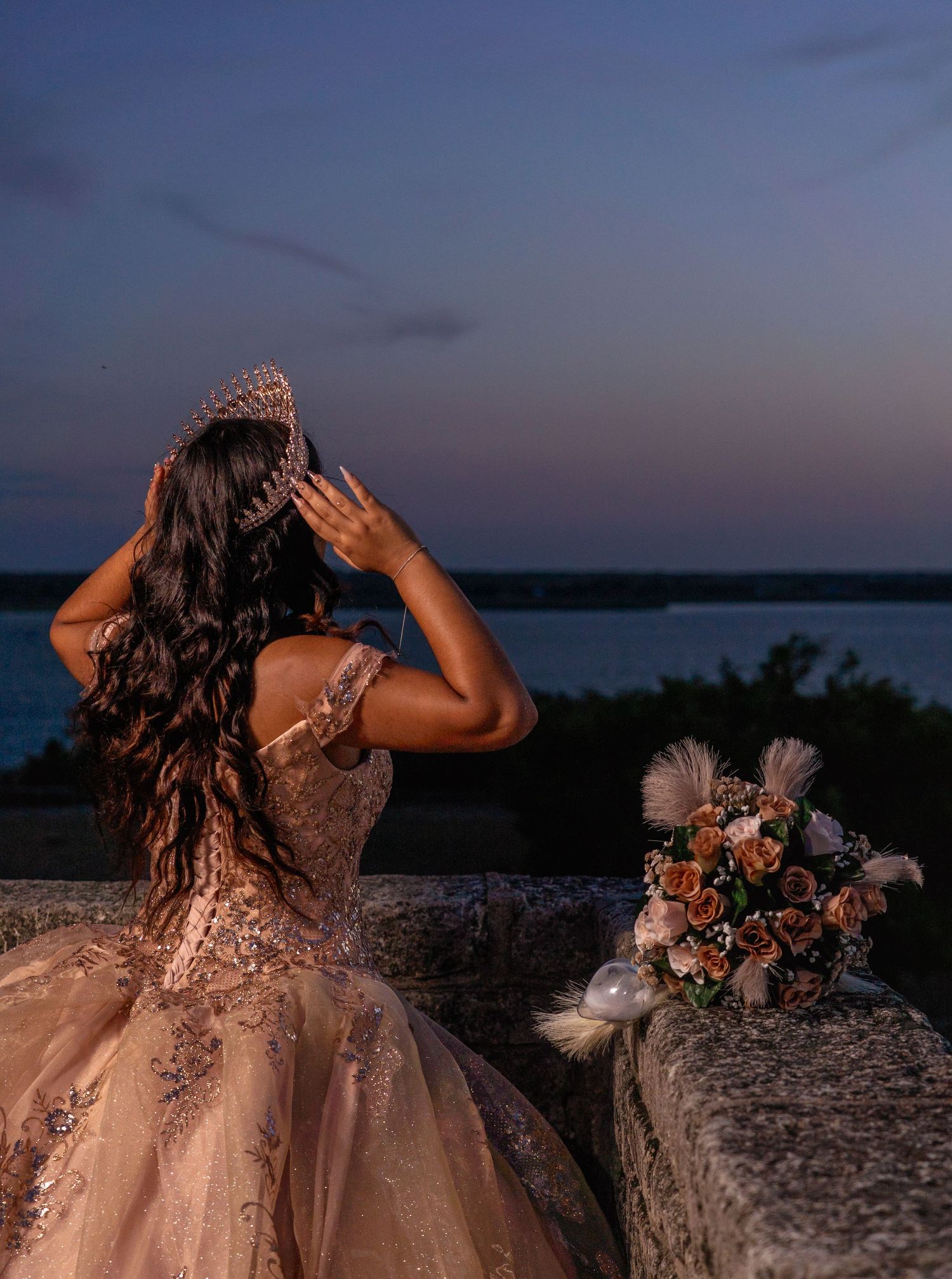 Figure in a sparkly ballgown gazes at sunset over water with bouquet on stone ledge in romantic dusk setting.