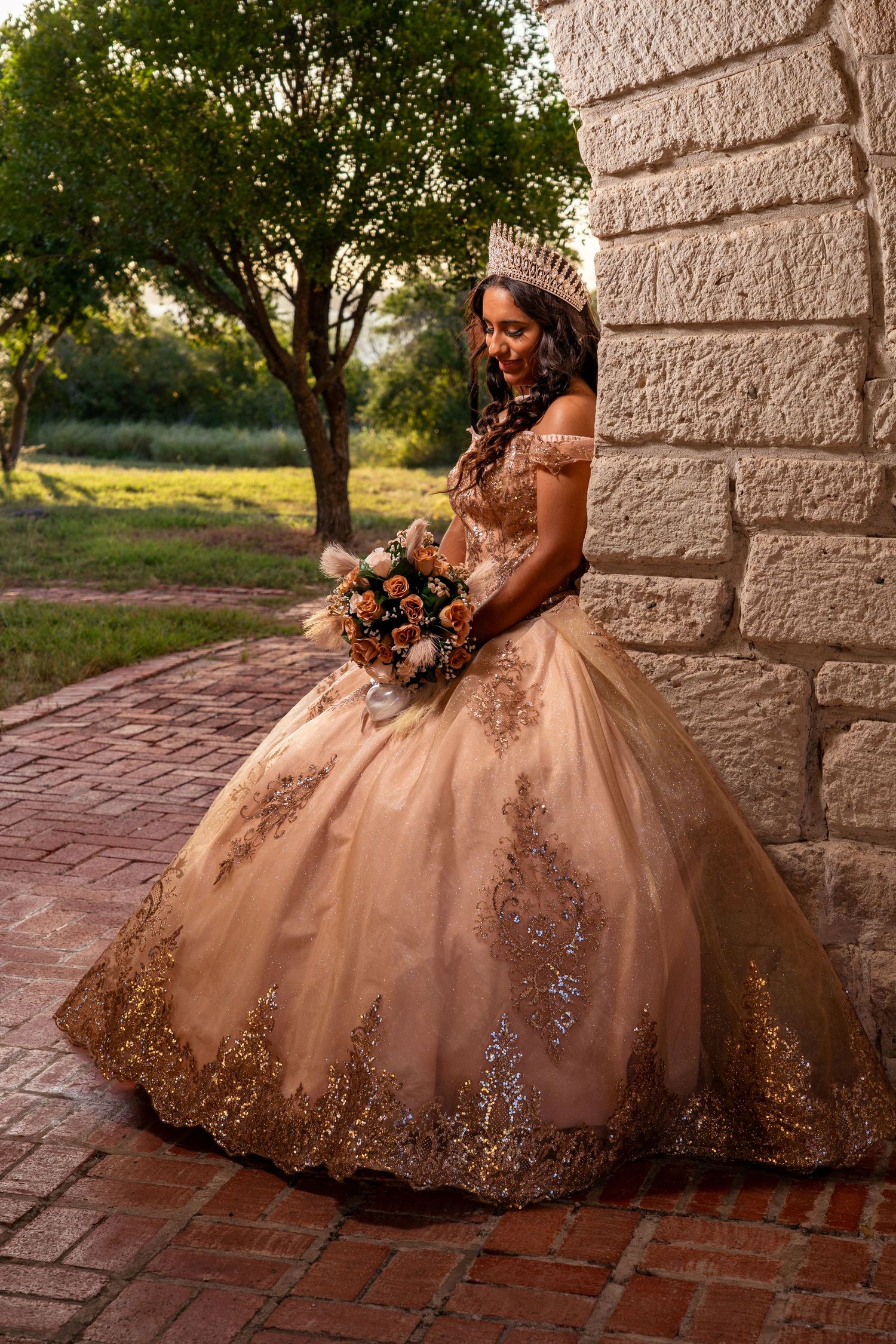An elegant rose gold quinceanera gown photographed against stone walls and trees at sunset.