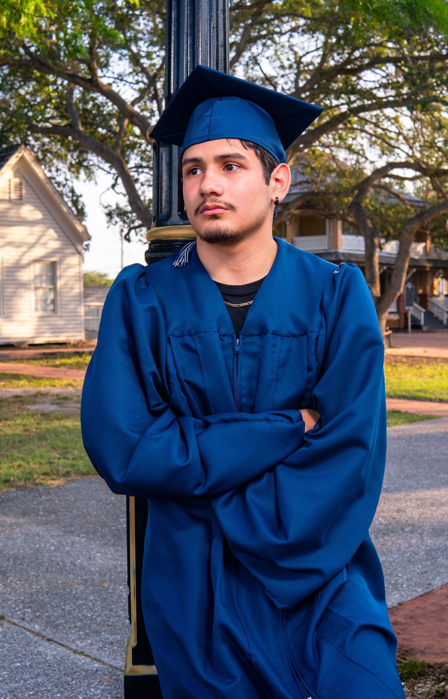 Graduate in blue cap and gown leans against lamp post on campus during golden hour.