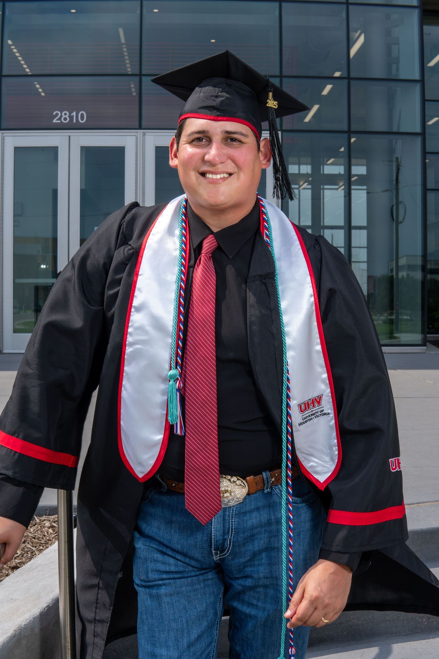 Graduate wearing black cap and gown with red accents, white stole, and red tie stands outside glass building.