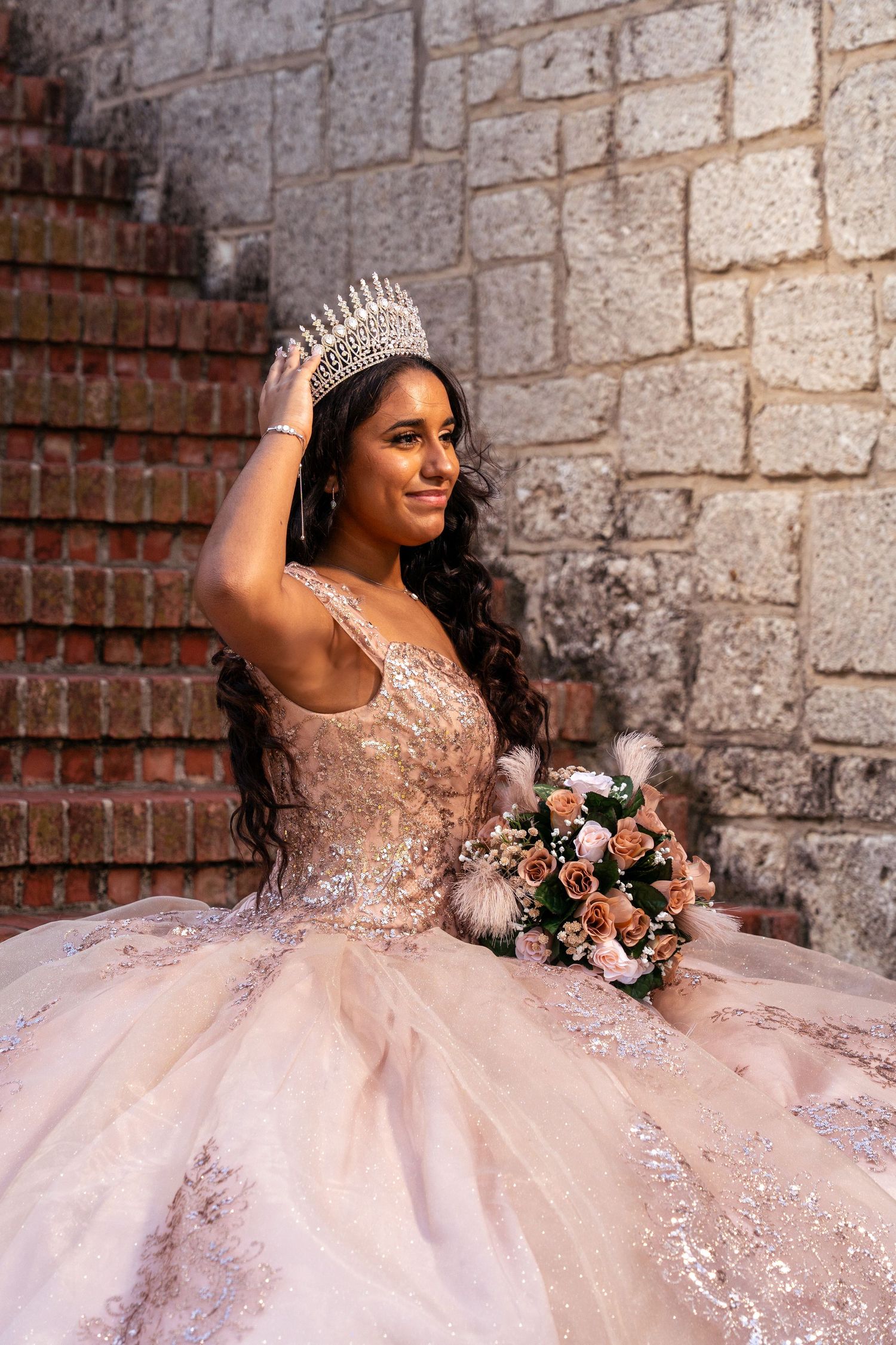 A person in a pink embellished ball gown with crown holding a blush and coral floral bouquet against a stone wall.