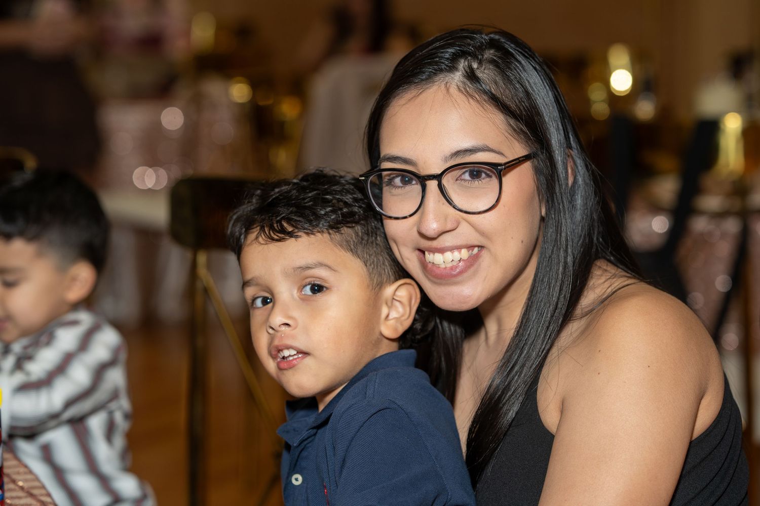 A smiling woman with glasses holds a young boy in a navy shirt at a festive family celebration.