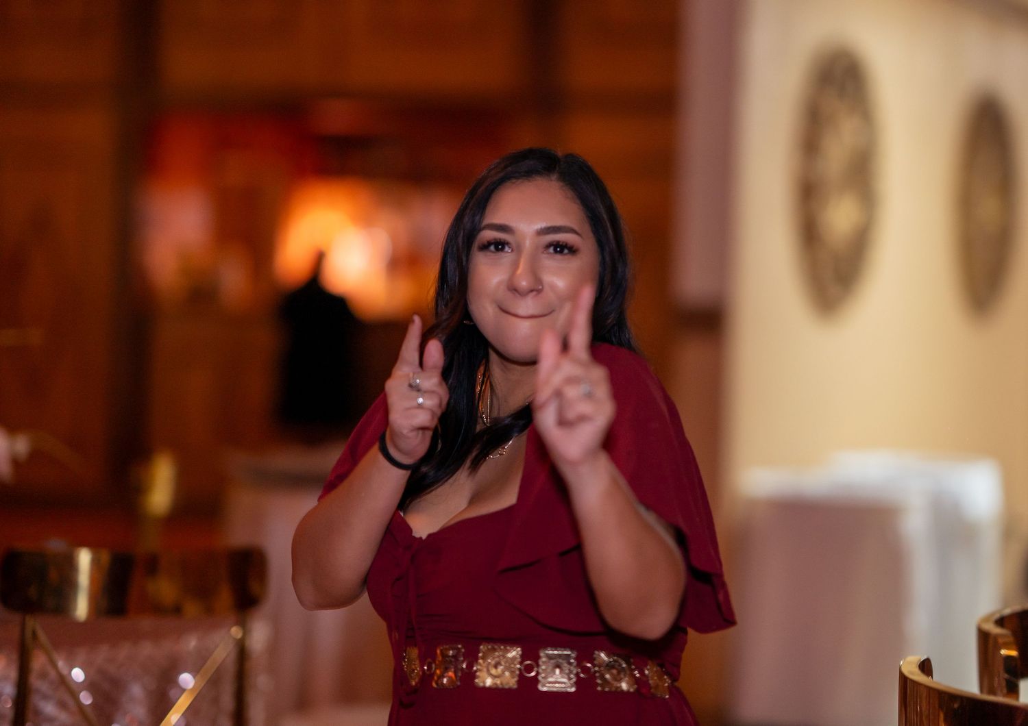 A woman in a burgundy dress with a gold belt smiles and gestures with both hands at an elegant event venue.