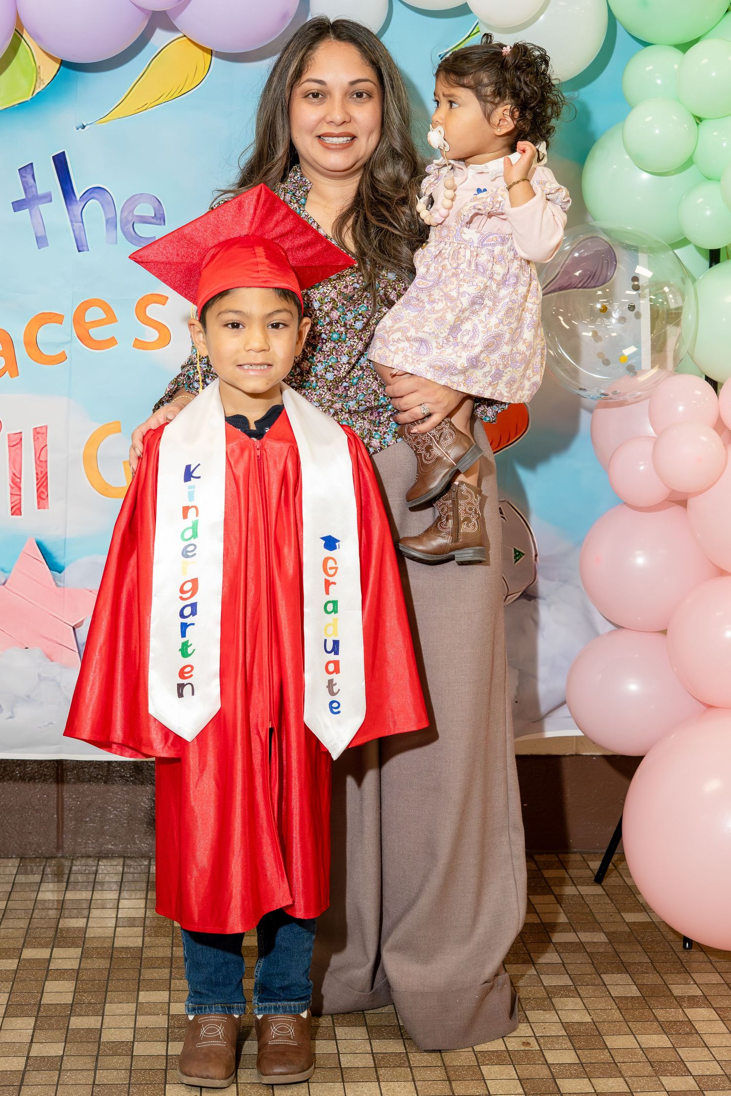 Young kindergarten graduate in red cap and gown poses with mother and baby sibling at graduation ceremony.