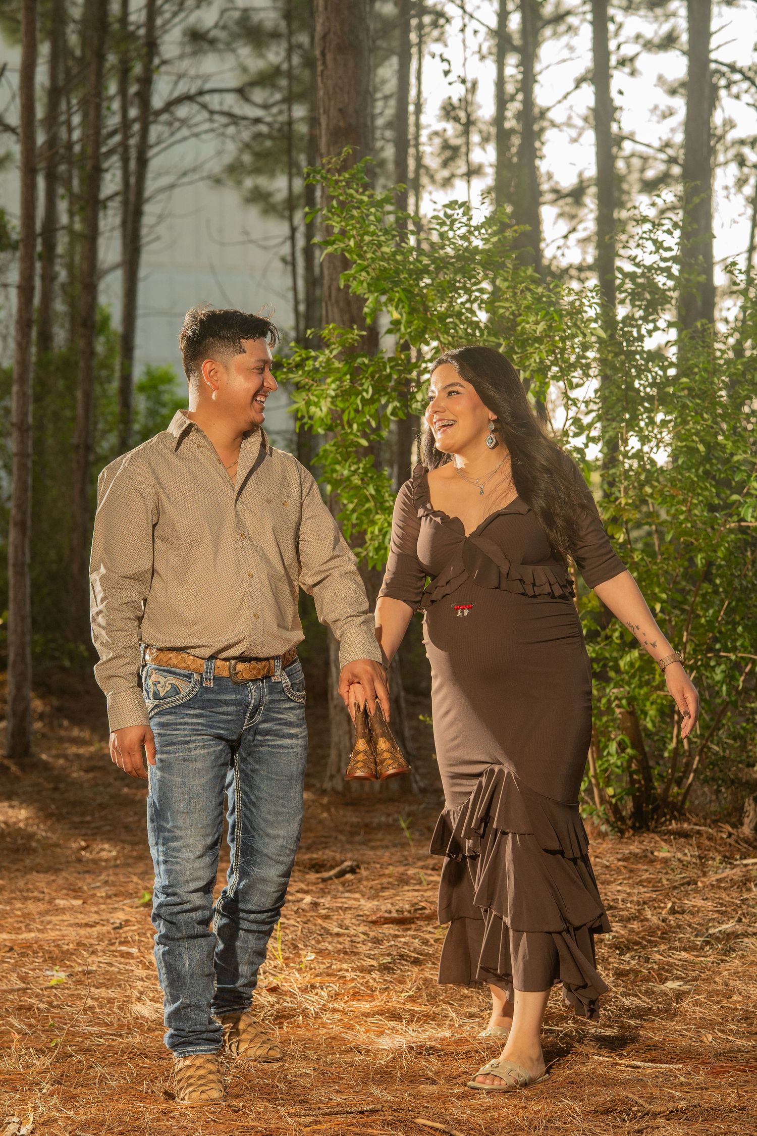 A smiling couple walks hand-in-hand through a pine forest, woman in brown dress, man in jeans and button-up shirt.