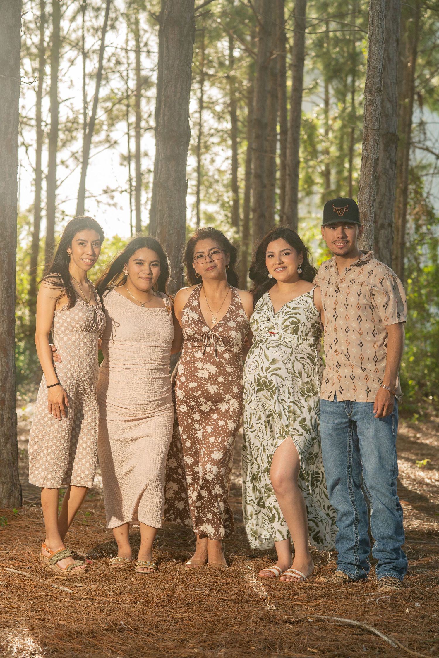 Family of five posing together in a sunlit pine forest, wearing casual summer outfits for an outdoor portrait.