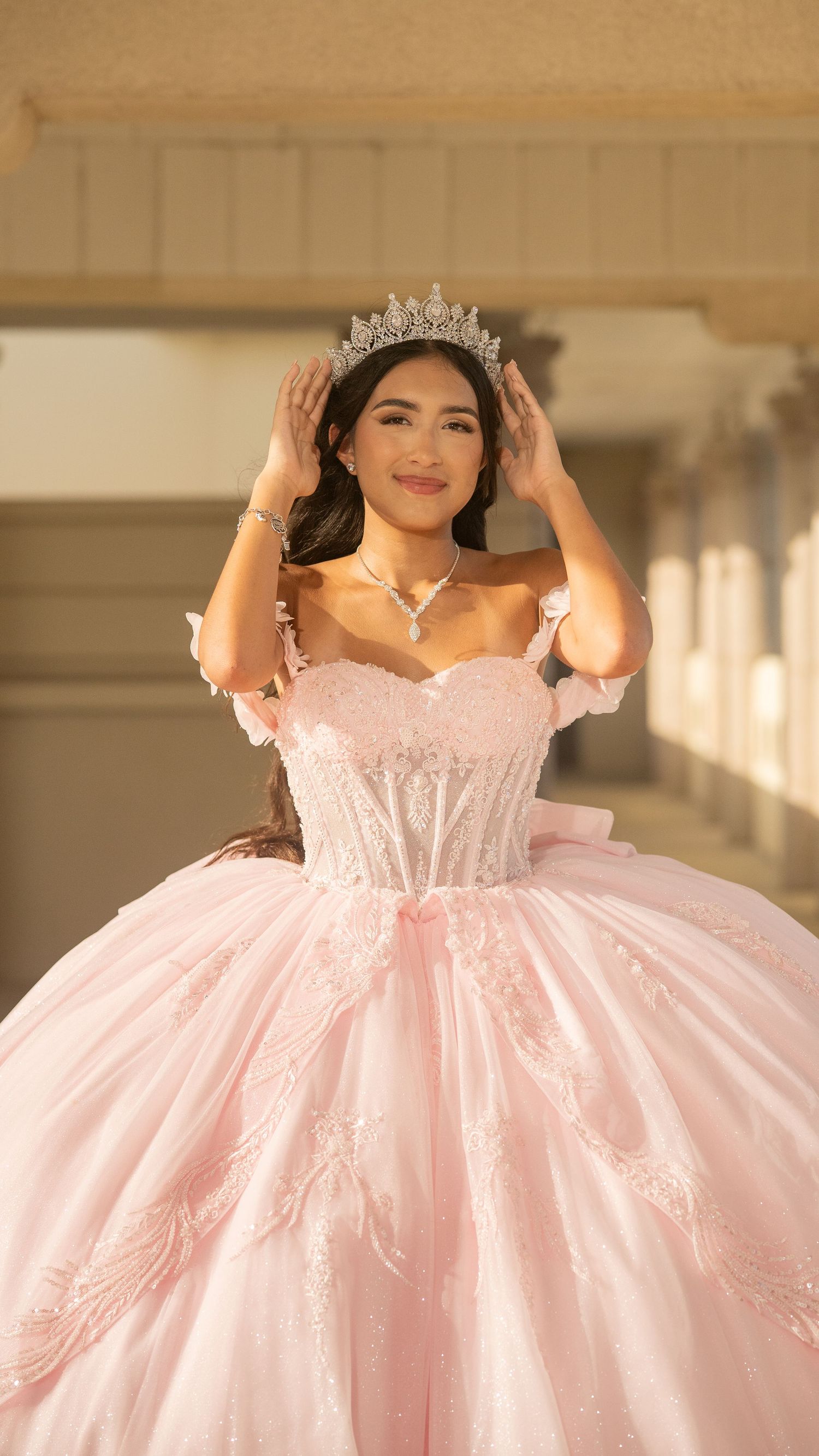 Young woman in pink quinceañera gown adjusts sparkling tiara crown in sunlit corridor.