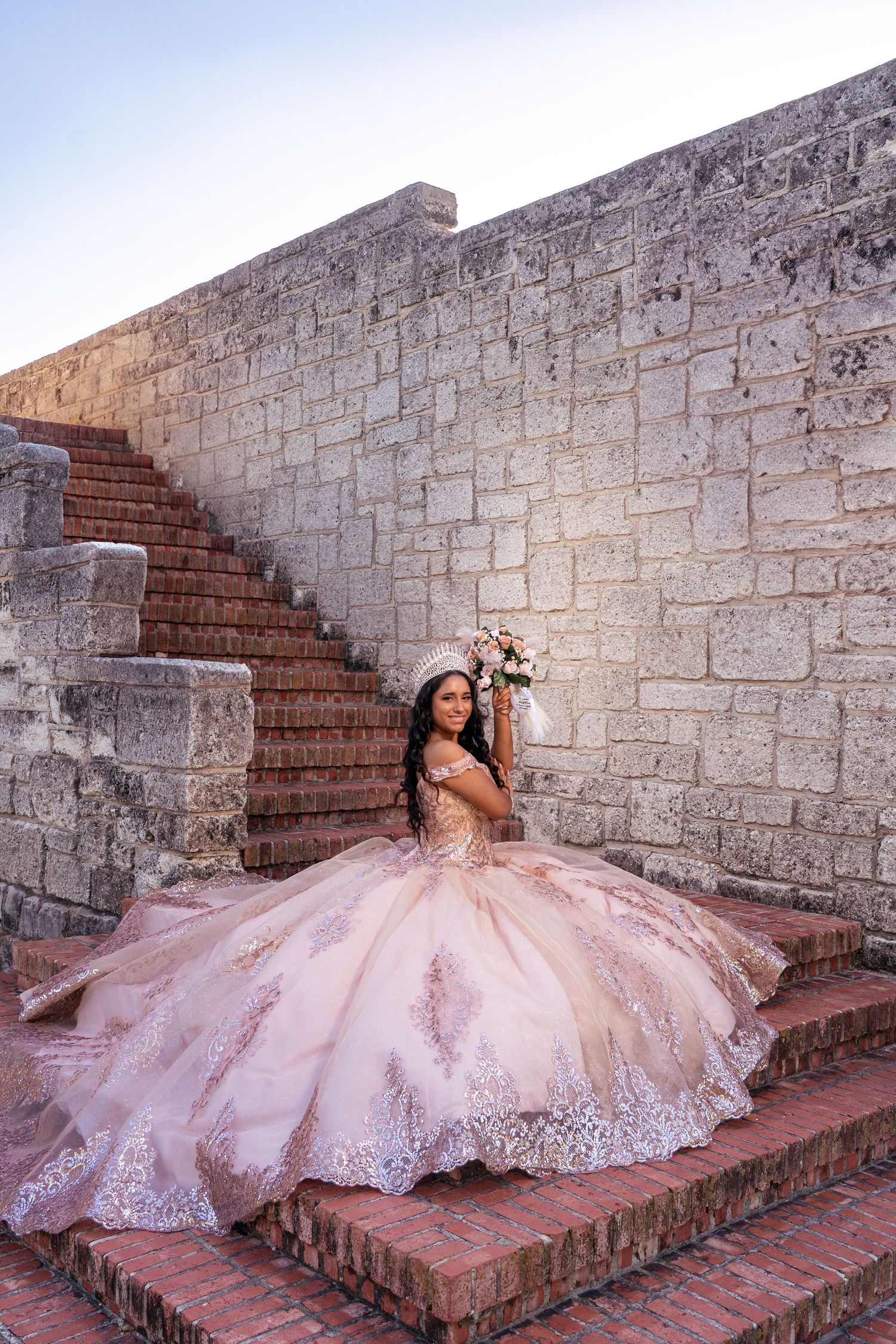 A blush pink ballgown with lace details photographed on stone steps outdoors.