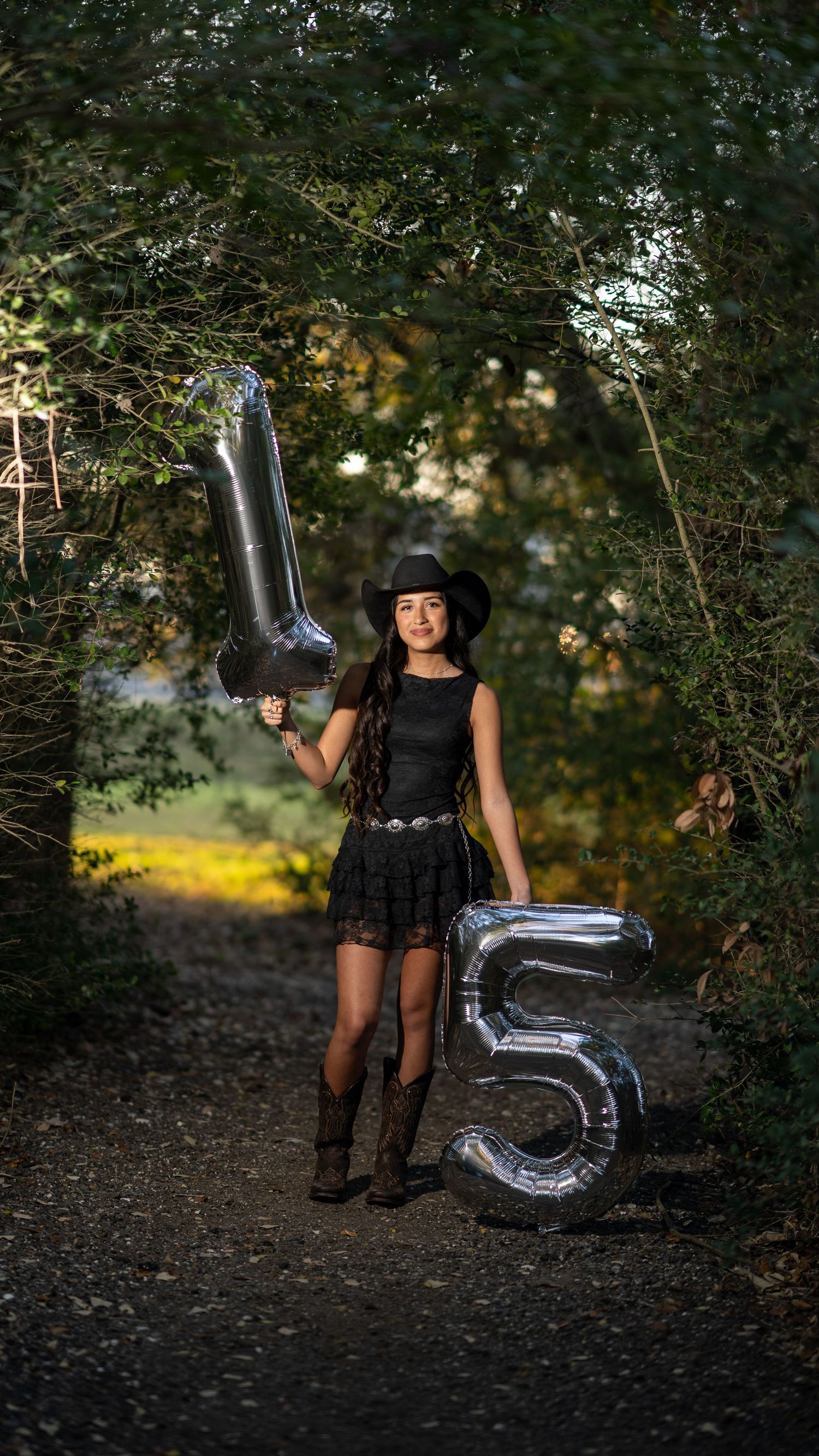 Young woman in black dress and cowboy hat posing with large silver 15 balloons on forest path.