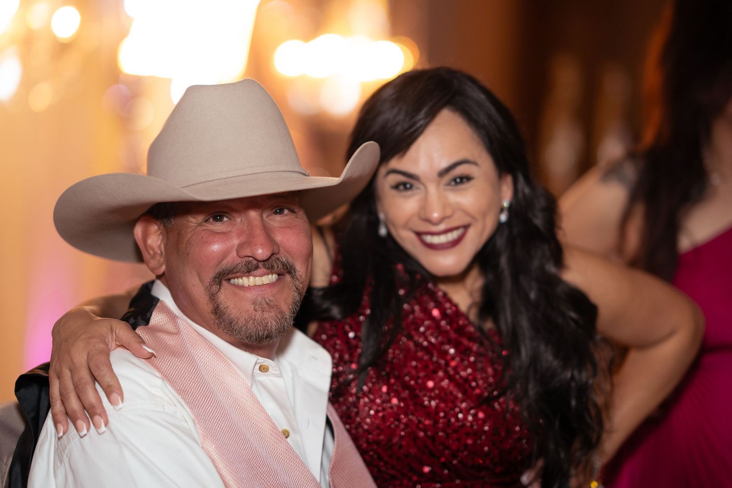 Man in beige cowboy hat smiles with woman in red sequin dress at a festive quinceañera celebration.