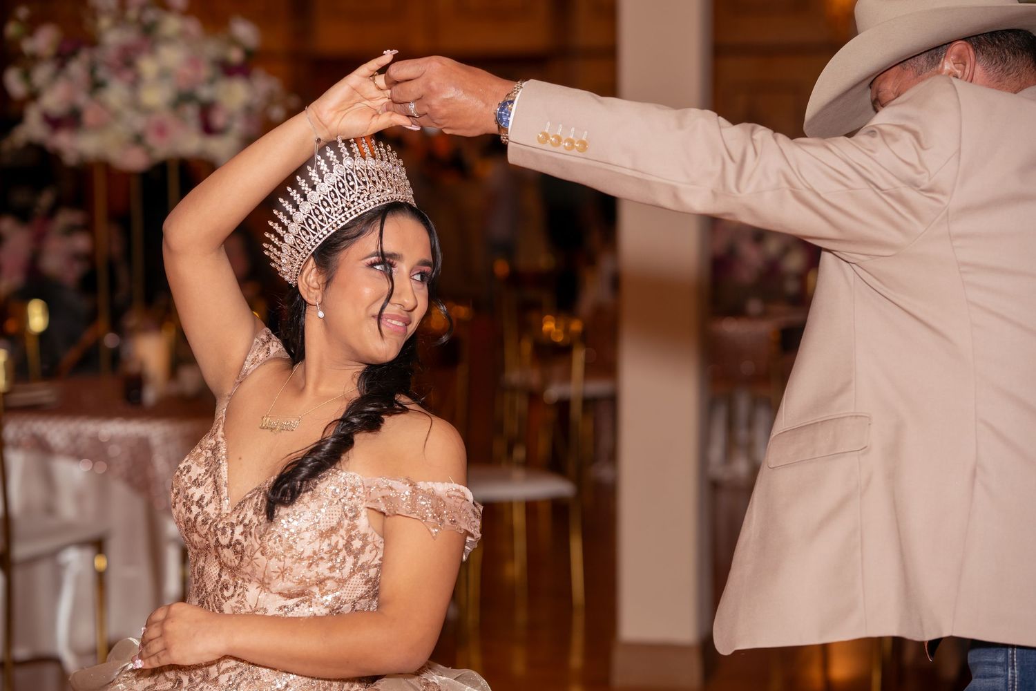 A man in a tan cowboy hat places a sparkling crown on a smiling quinceañera girl in a rose gold gown.