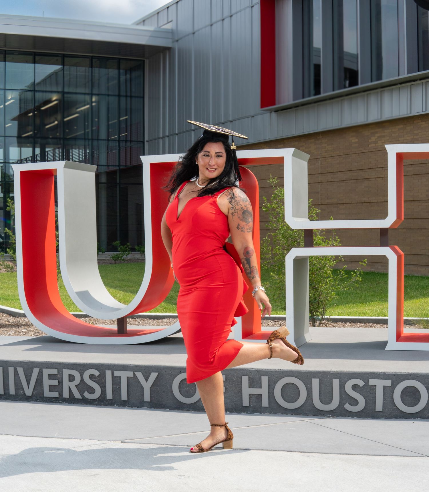 Graduate celebrates by University of Houston UH sign in red dress on campus grounds.
