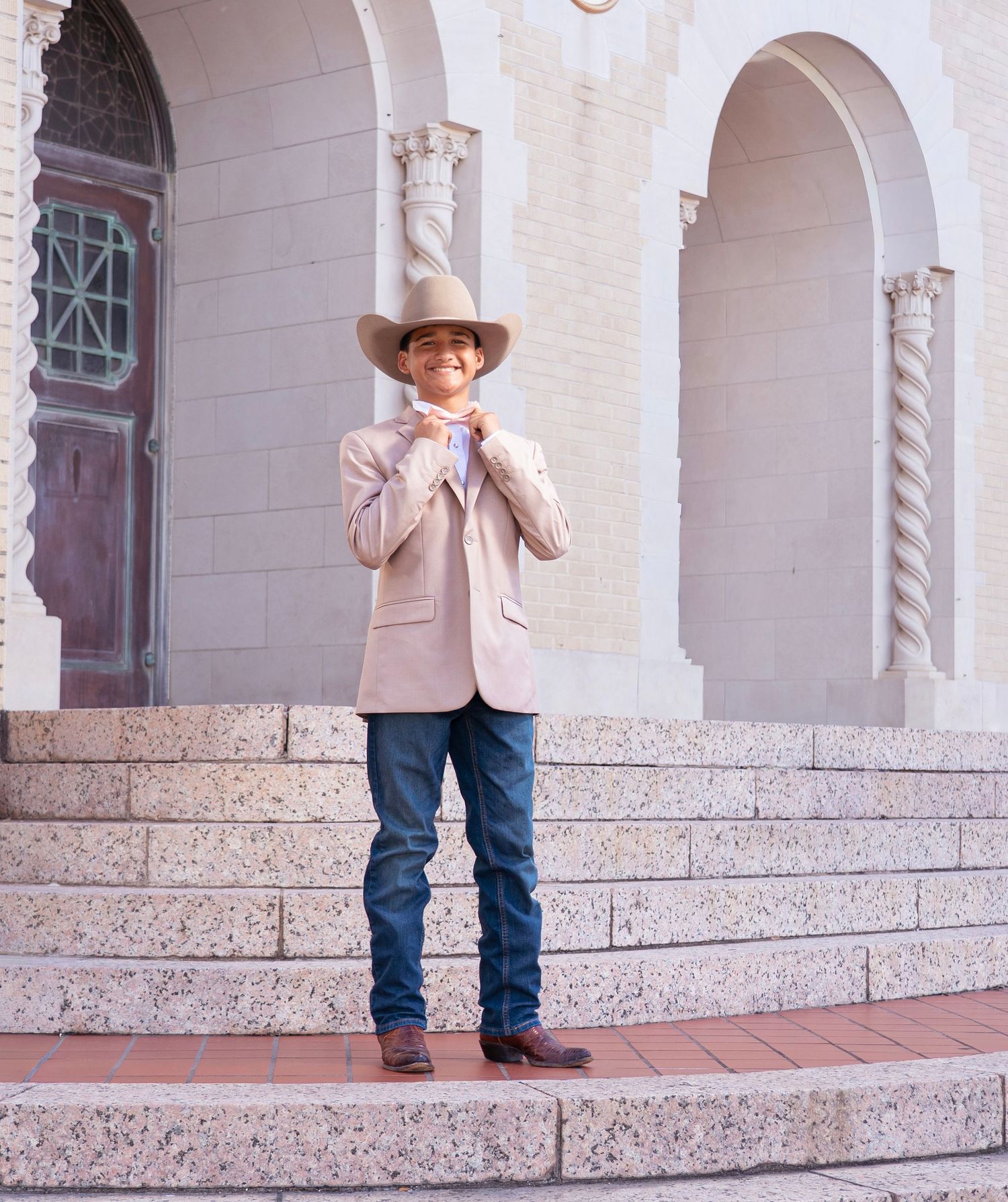 Person in cowboy hat and jeans stands on ornate stone steps of historic building with arched columns.
