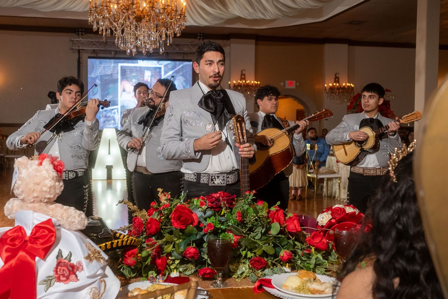 A mariachi band performs at an elegant event with red roses and a crystal chandelier in the background.