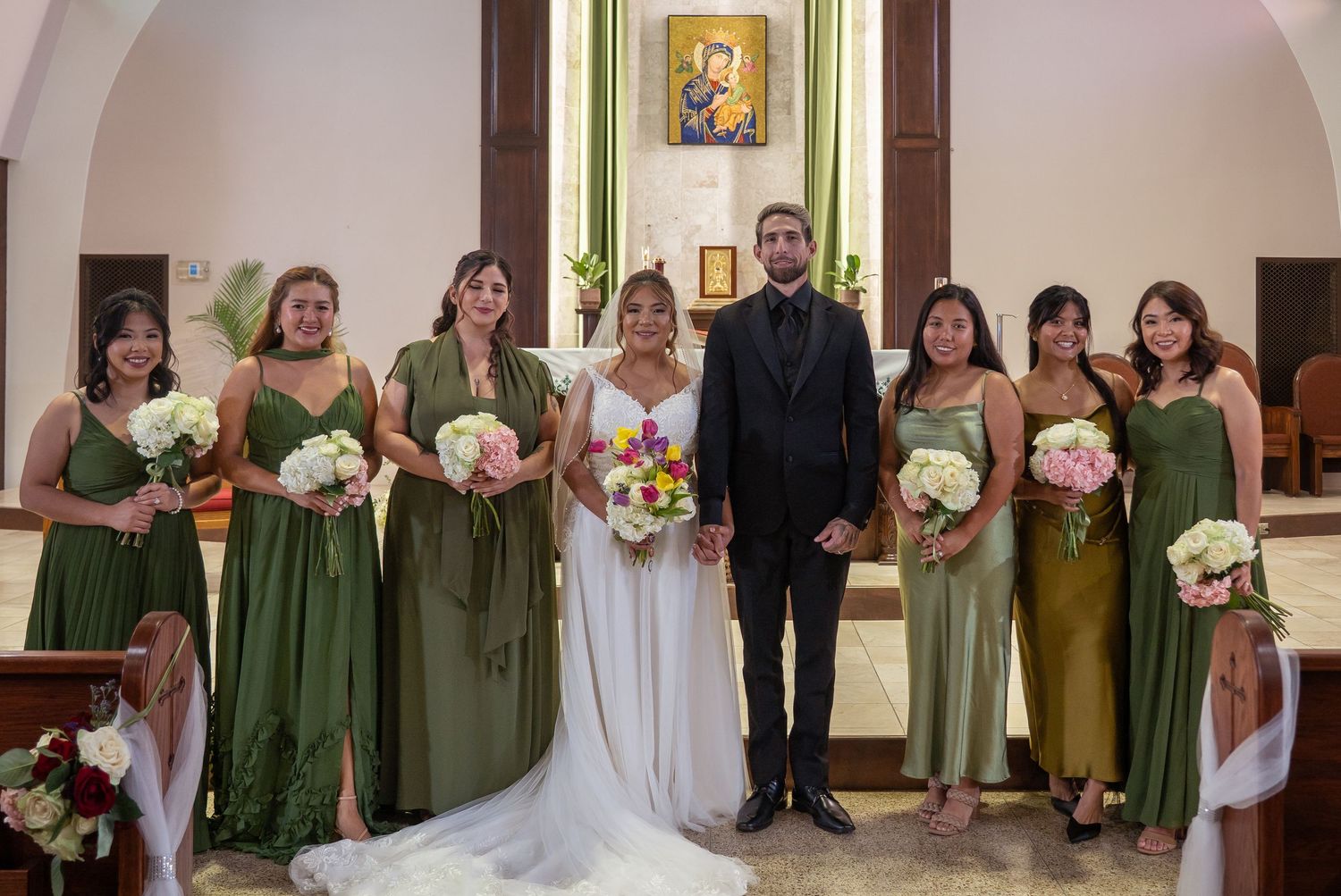 Wedding party posing in church, bride in white gown with groom in black suit, flanked by six bridesmaids in olive green dresses.