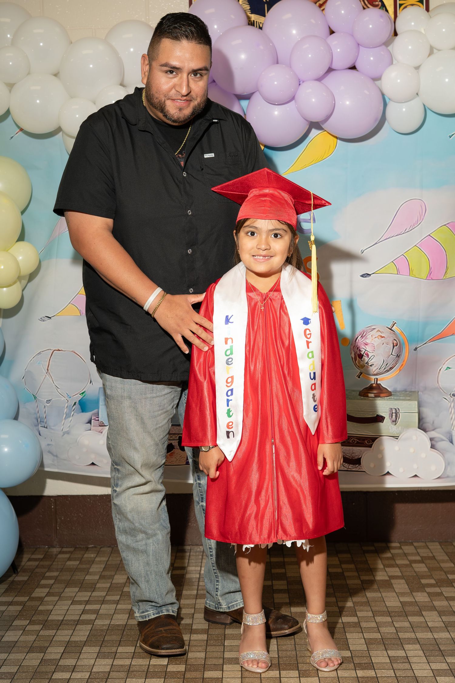 A young girl in a red graduation gown and cap poses with a man in a black shirt at a kindergarten graduation ceremony.