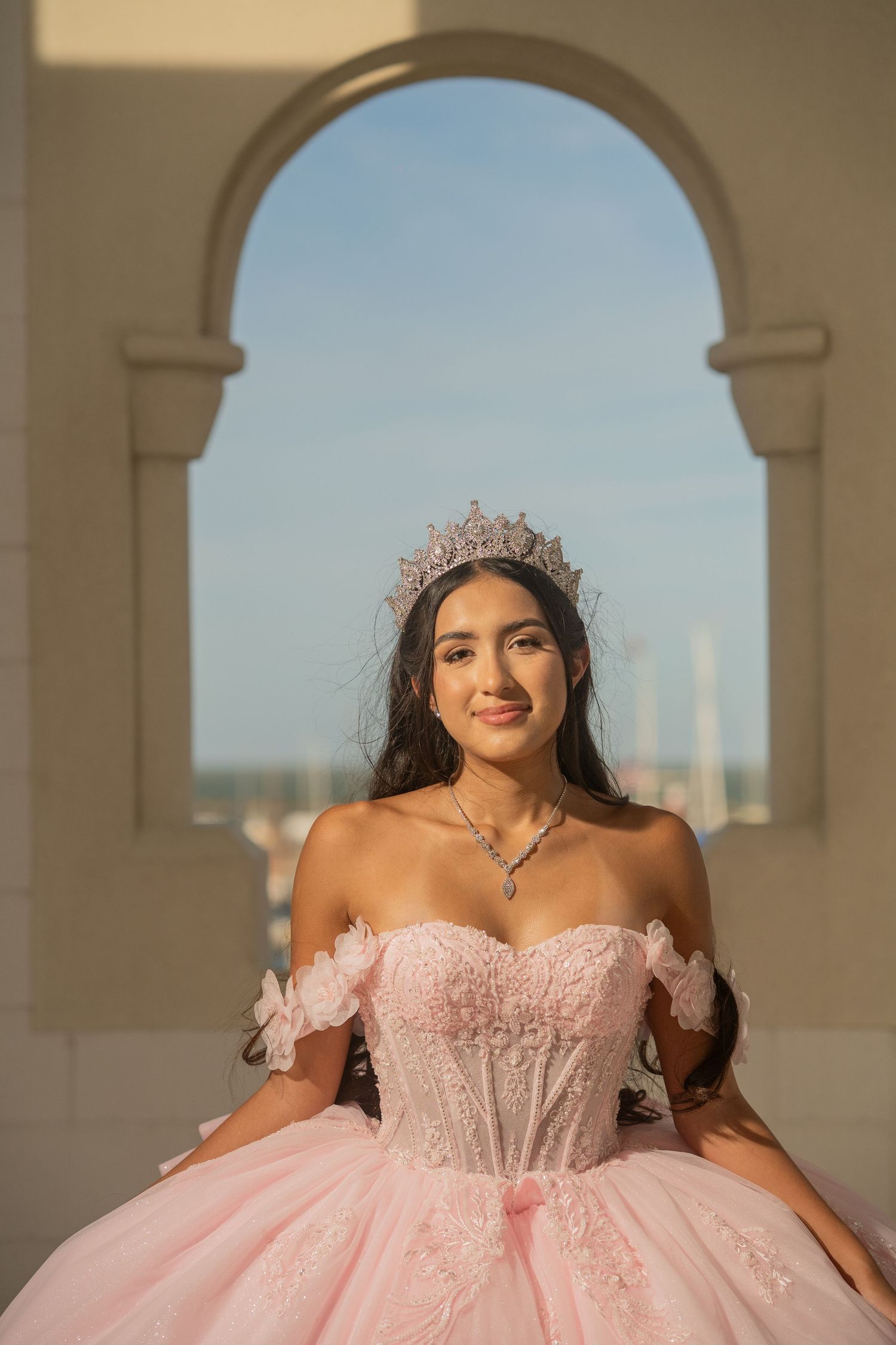 Young woman in pink quinceañera gown and crystal crown poses gracefully under an archway with blue sky backdrop.