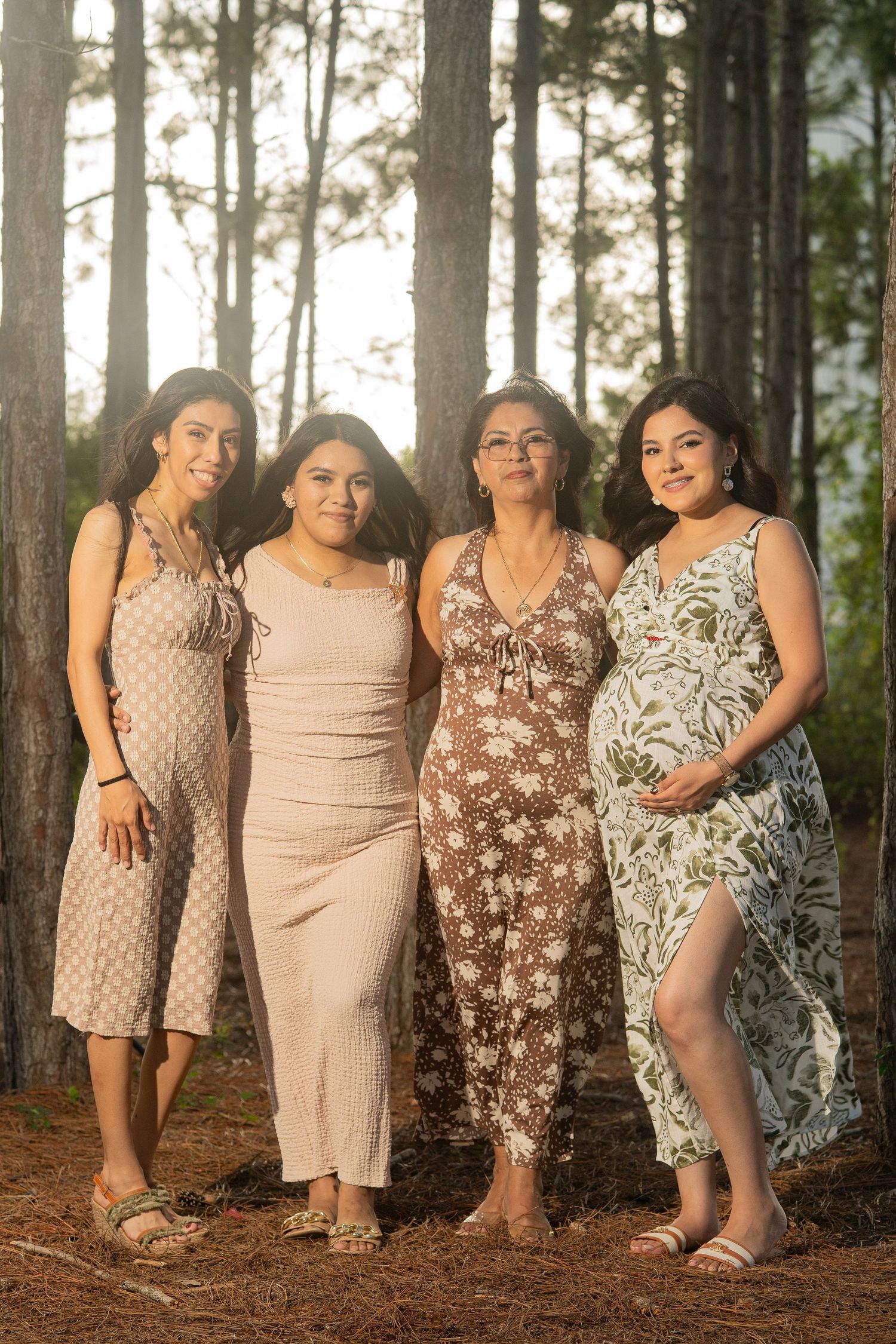 Four women in summer dresses pose together in a pine forest, one visibly pregnant, smiling for a family portrait.