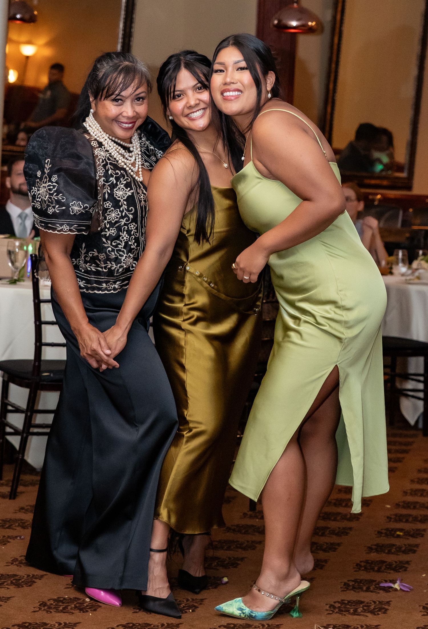 Three women smile together in elegant dresses at a wedding reception dinner.