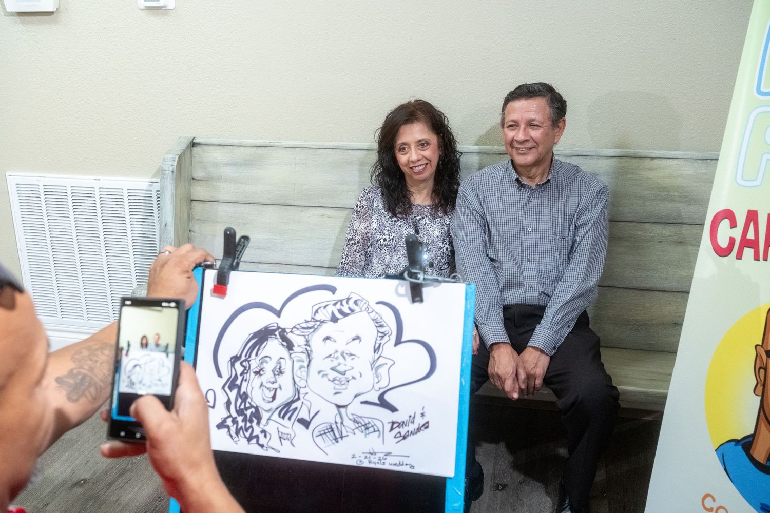 A couple poses beside a caricature drawing while a photographer captures the moment at an indoor event.