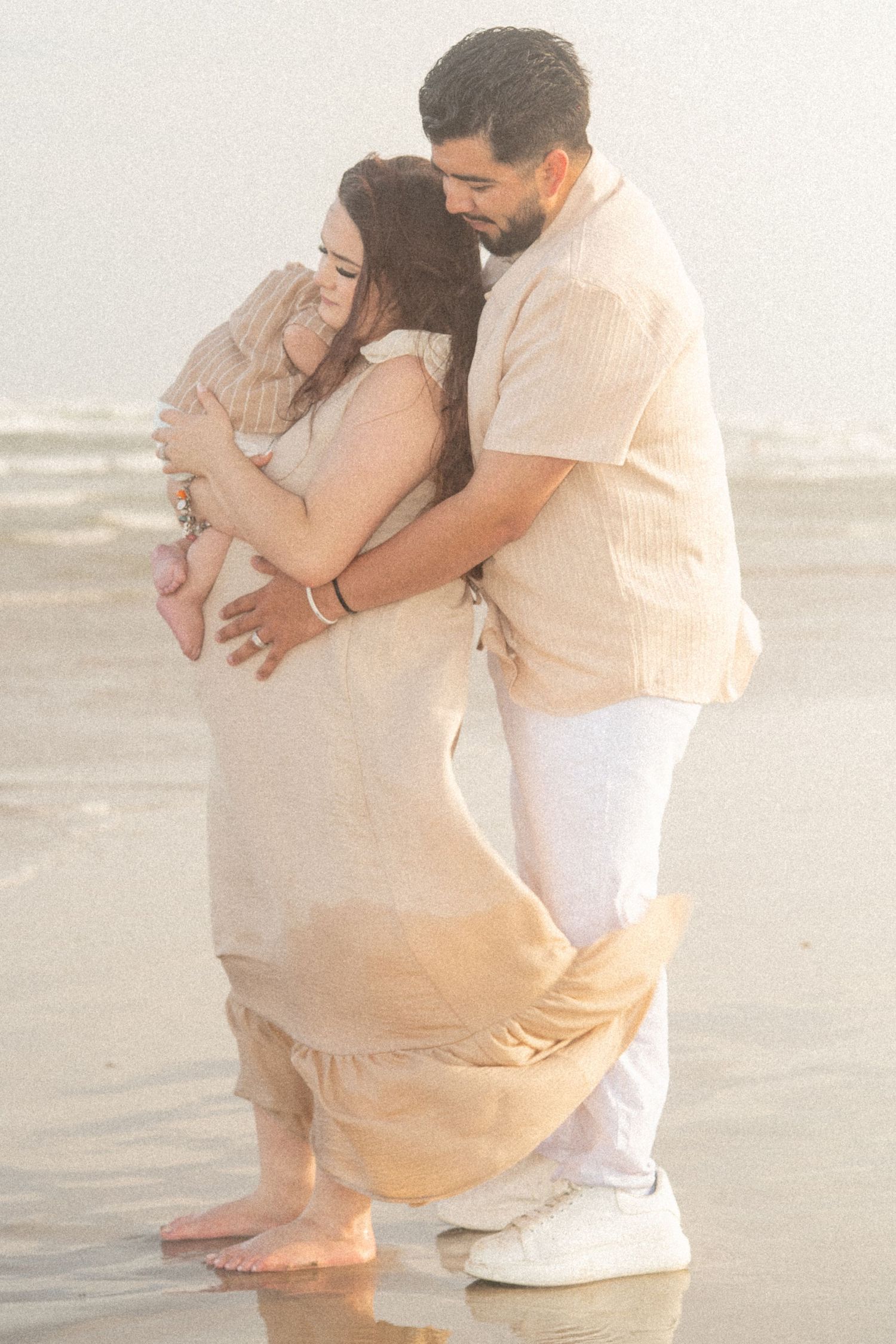 Couple shares a tender embrace on a misty beach, woman in flowing beige dress held by bearded partner.