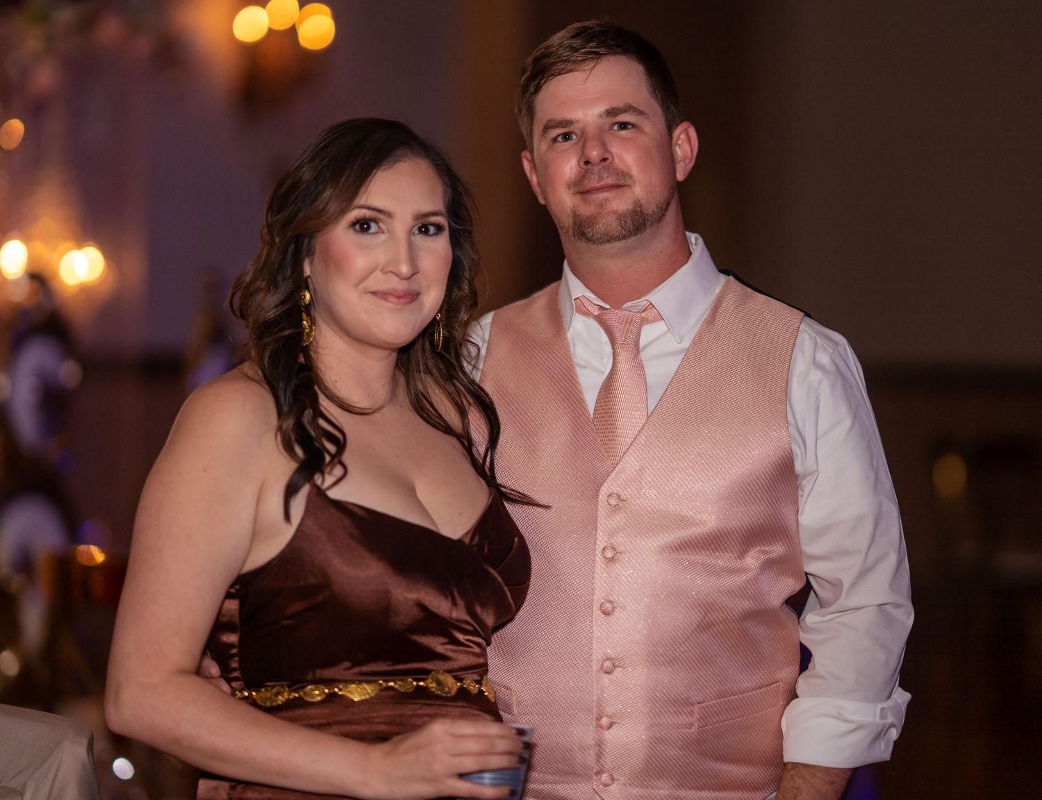 Couple posing together at formal event, woman in brown satin gown, man in pink vest and tie.