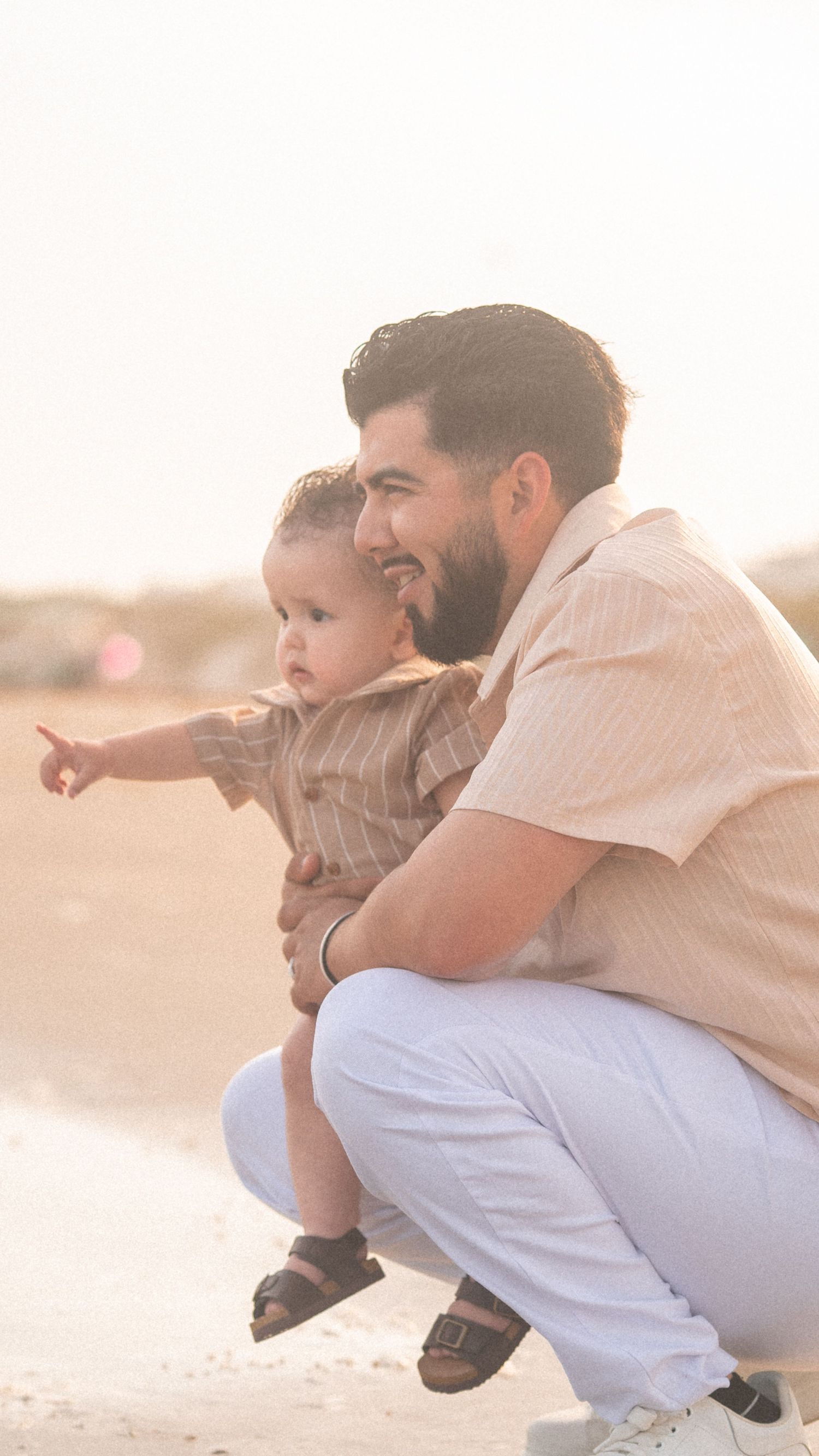 A smiling father crouches on a beach holding his baby who points toward the ocean.