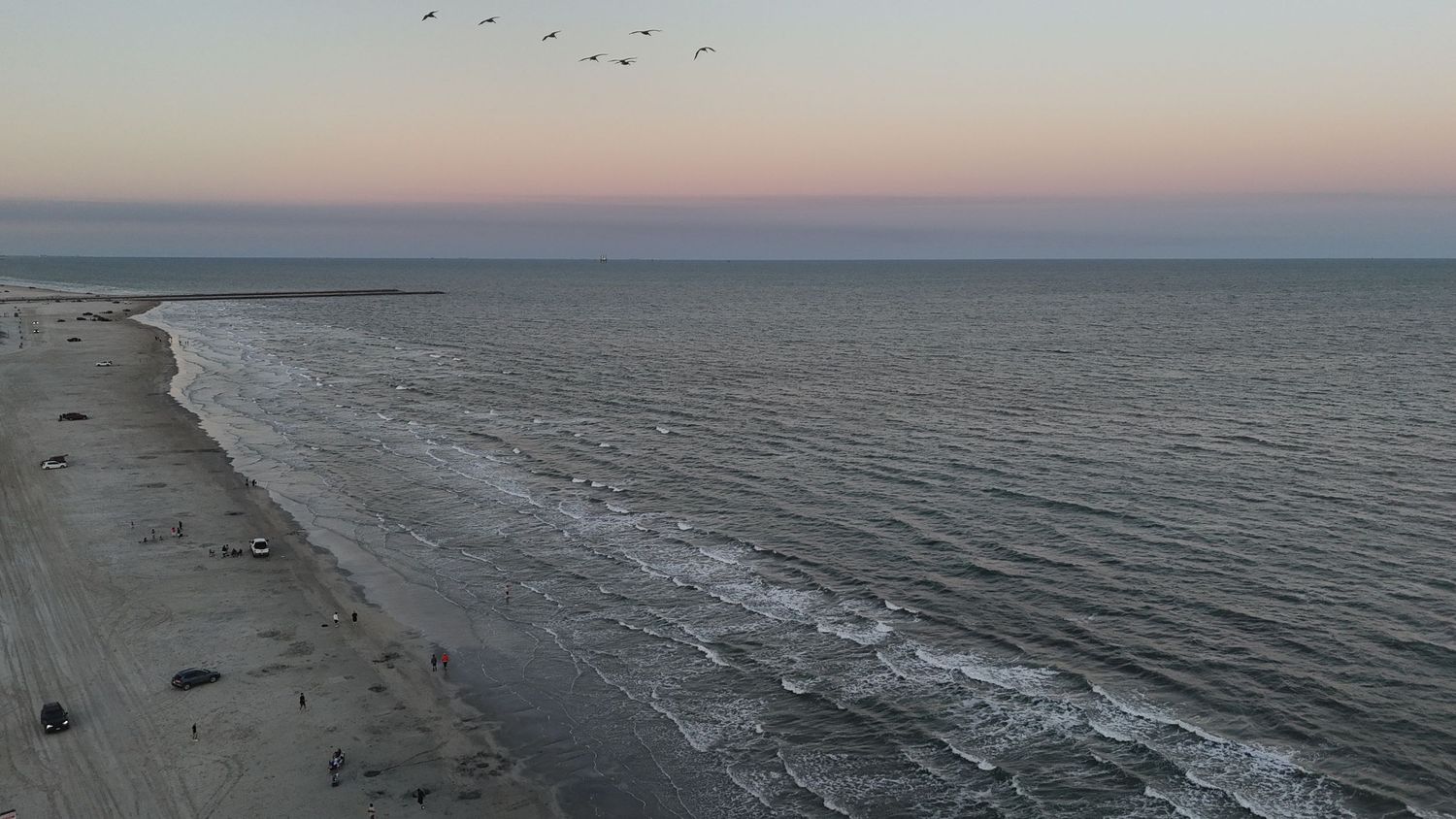 Aerial drone view of a peaceful beach coastline at dusk with birds flying over the ocean waves.