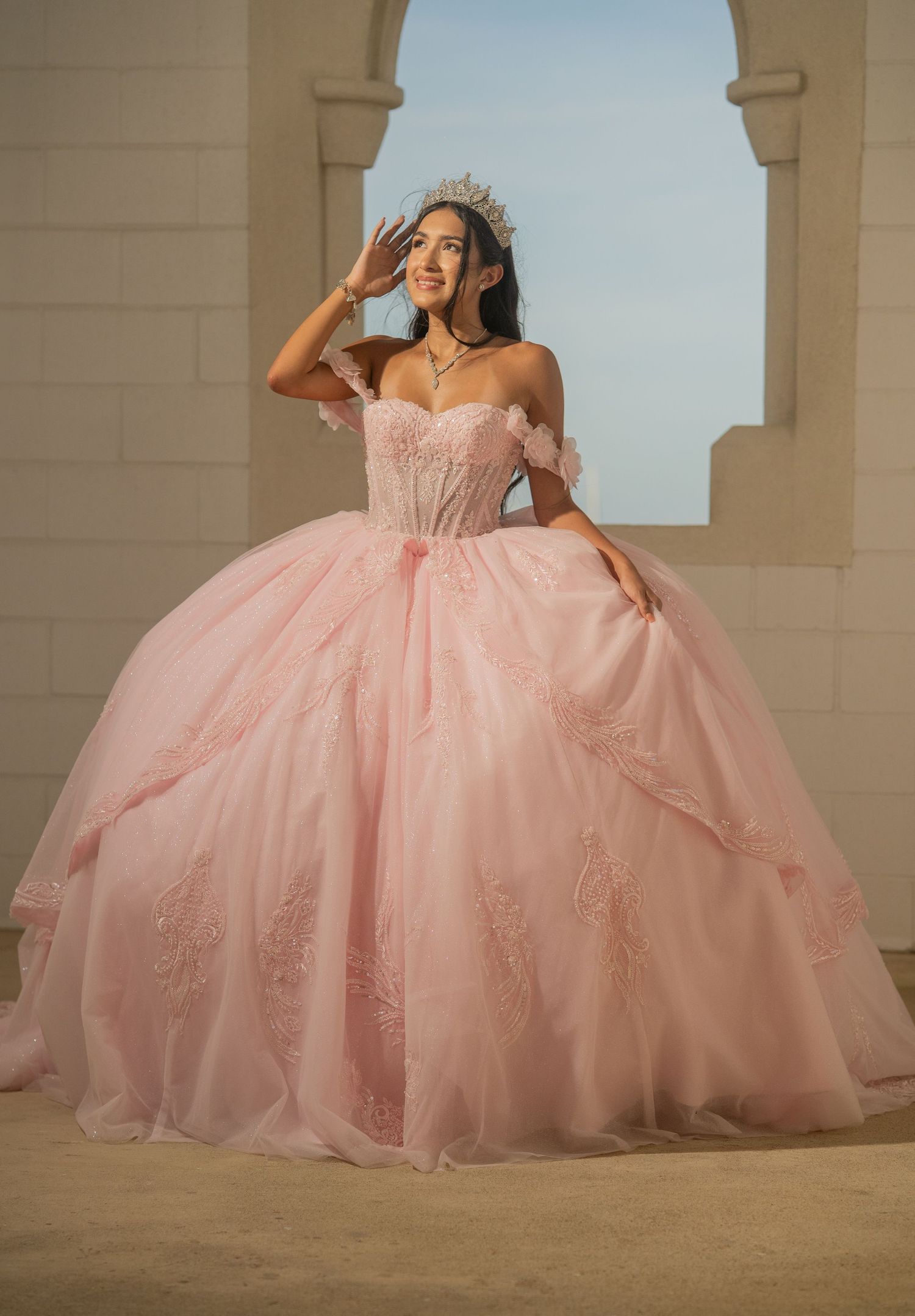 Woman wearing a pink quinceañera ball gown with floral details and tiara poses outdoors.
