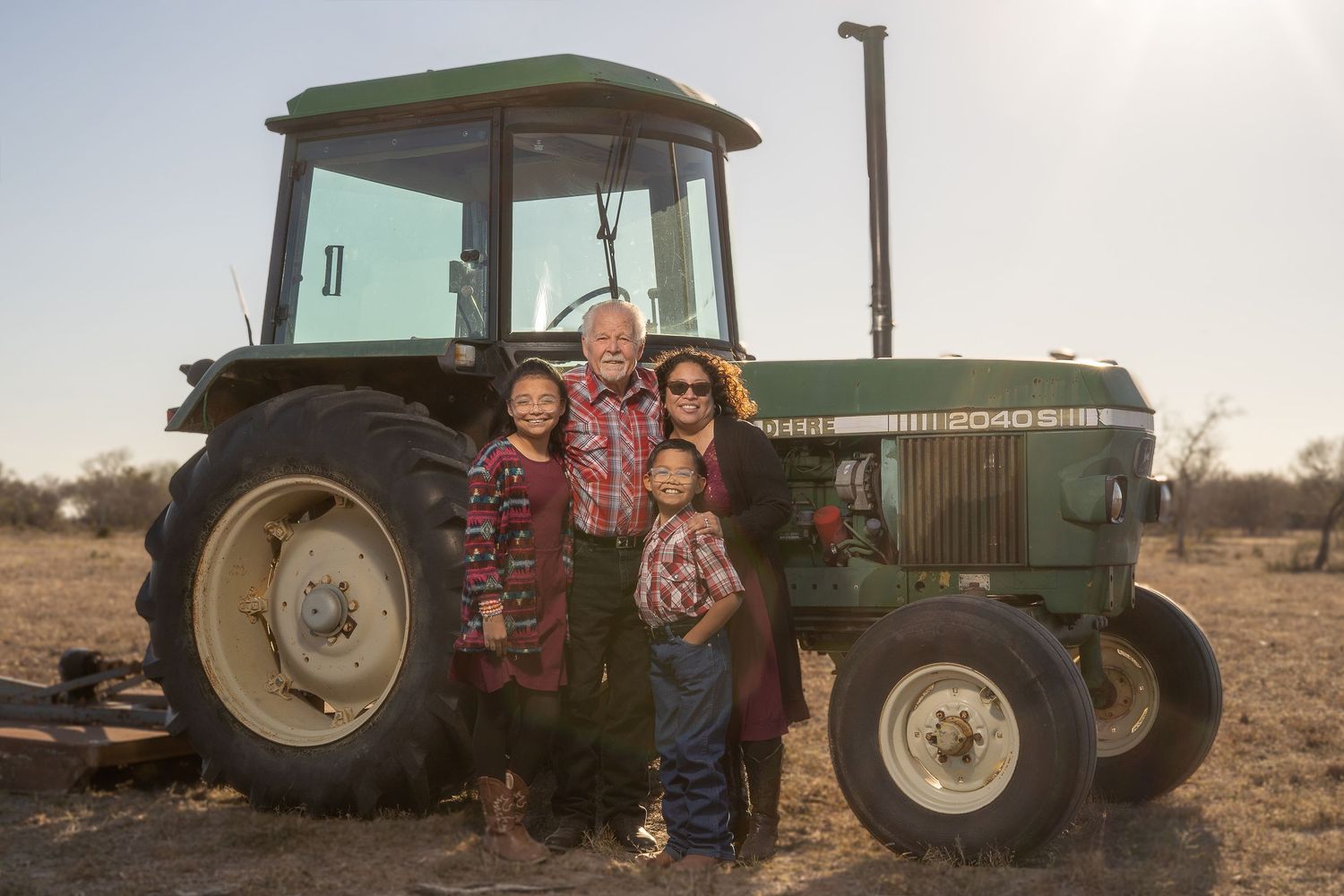 Family of four posing in front of a green John Deere tractor on a farm at sunset.