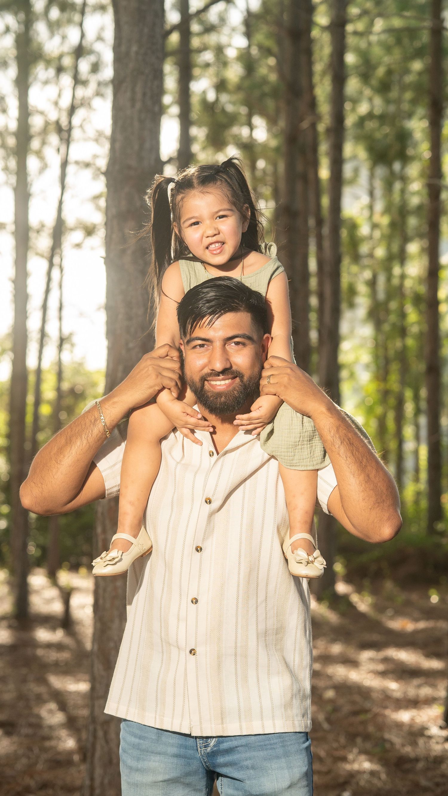 Smiling bearded man carries a happy toddler girl on his shoulders in a sunny pine forest.
