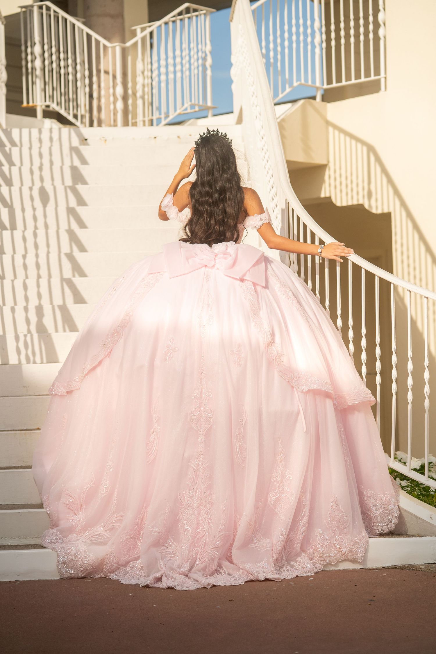 Young woman in a blush pink ball gown with lace details and bow, ascending white stairs wearing a tiara.