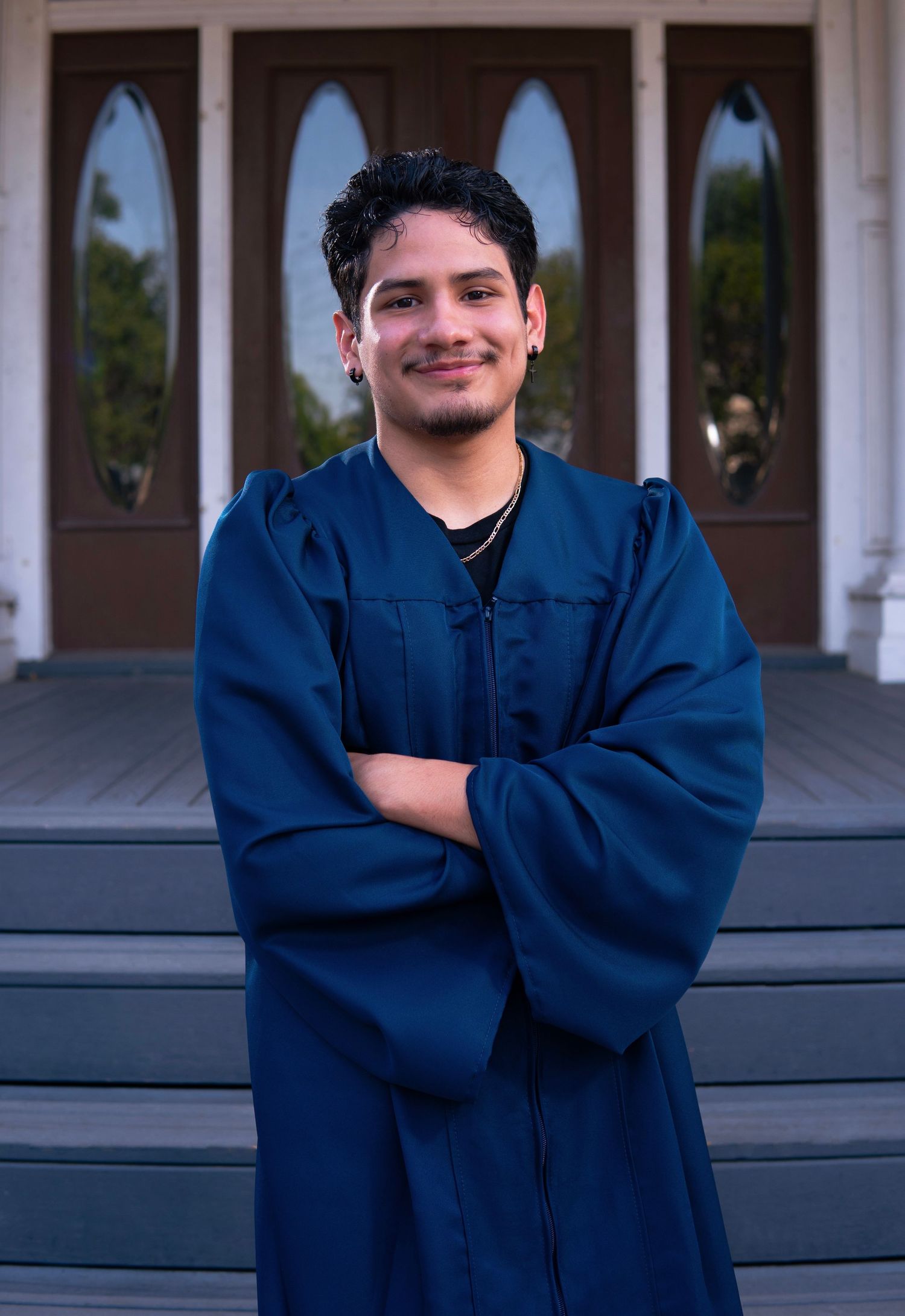 Graduate in navy blue graduation gown stands smiling with arms crossed in front of wooden doors.