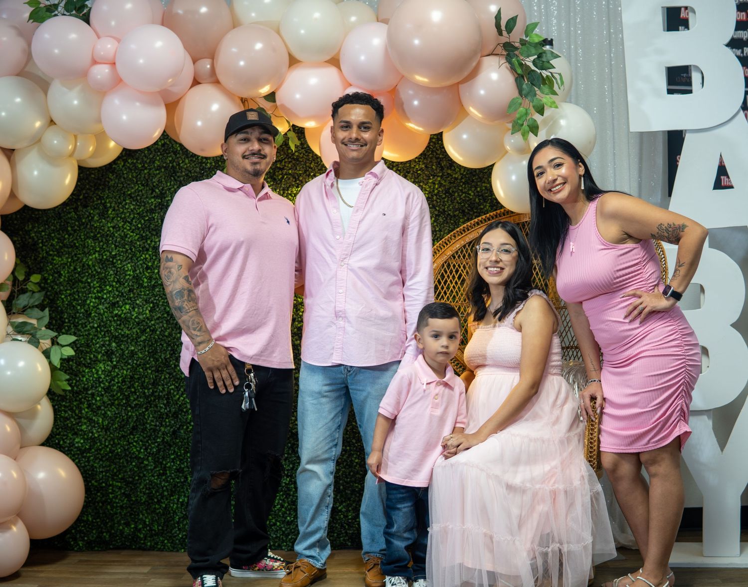 Family wearing matching pink outfits poses together against balloon arch and greenery backdrop at baby shower celebration.