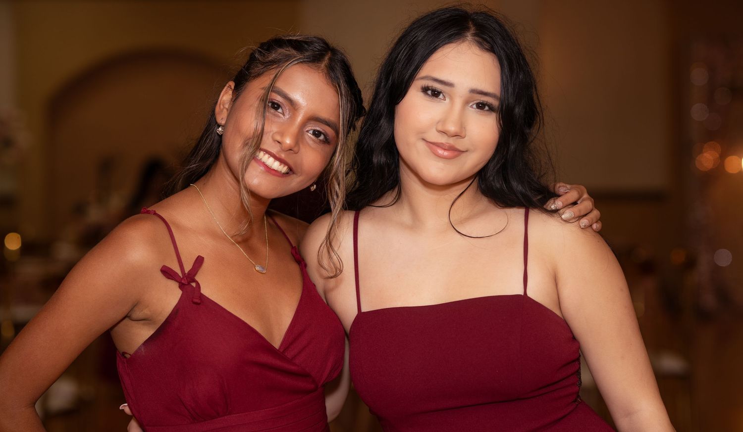 Two young women in matching burgundy dresses smile together at a formal quinceañera celebration.
