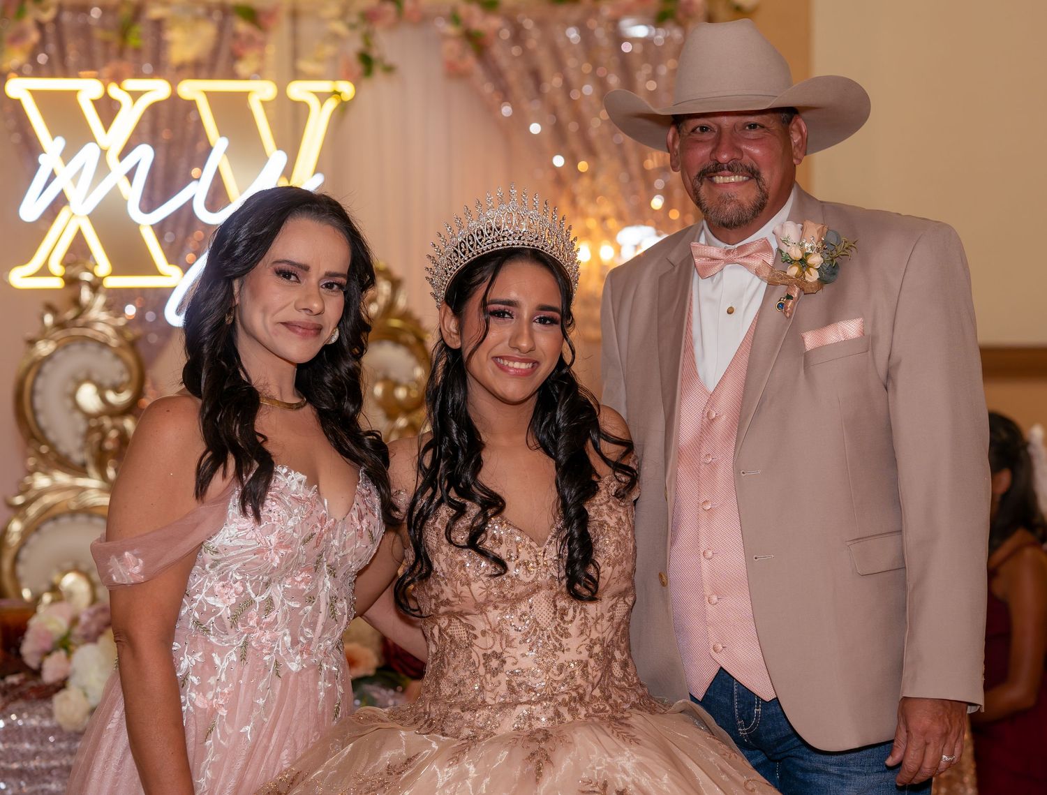 Quinceañera girl in gold gown and tiara poses with parents at her XV años celebration before a neon sign.