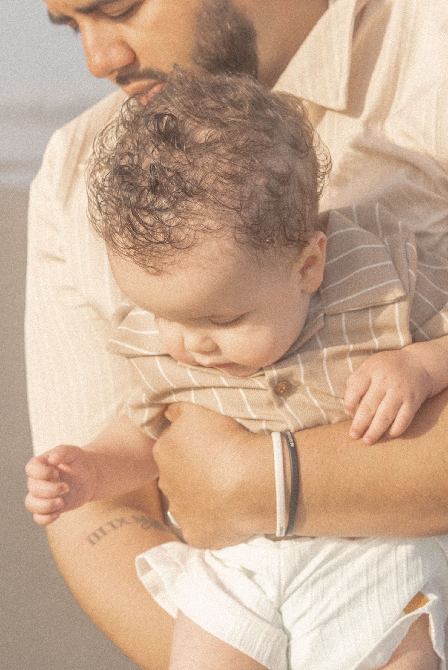 A father in a beige shirt gently cradles his sleeping curly-haired baby against his chest on a beach.