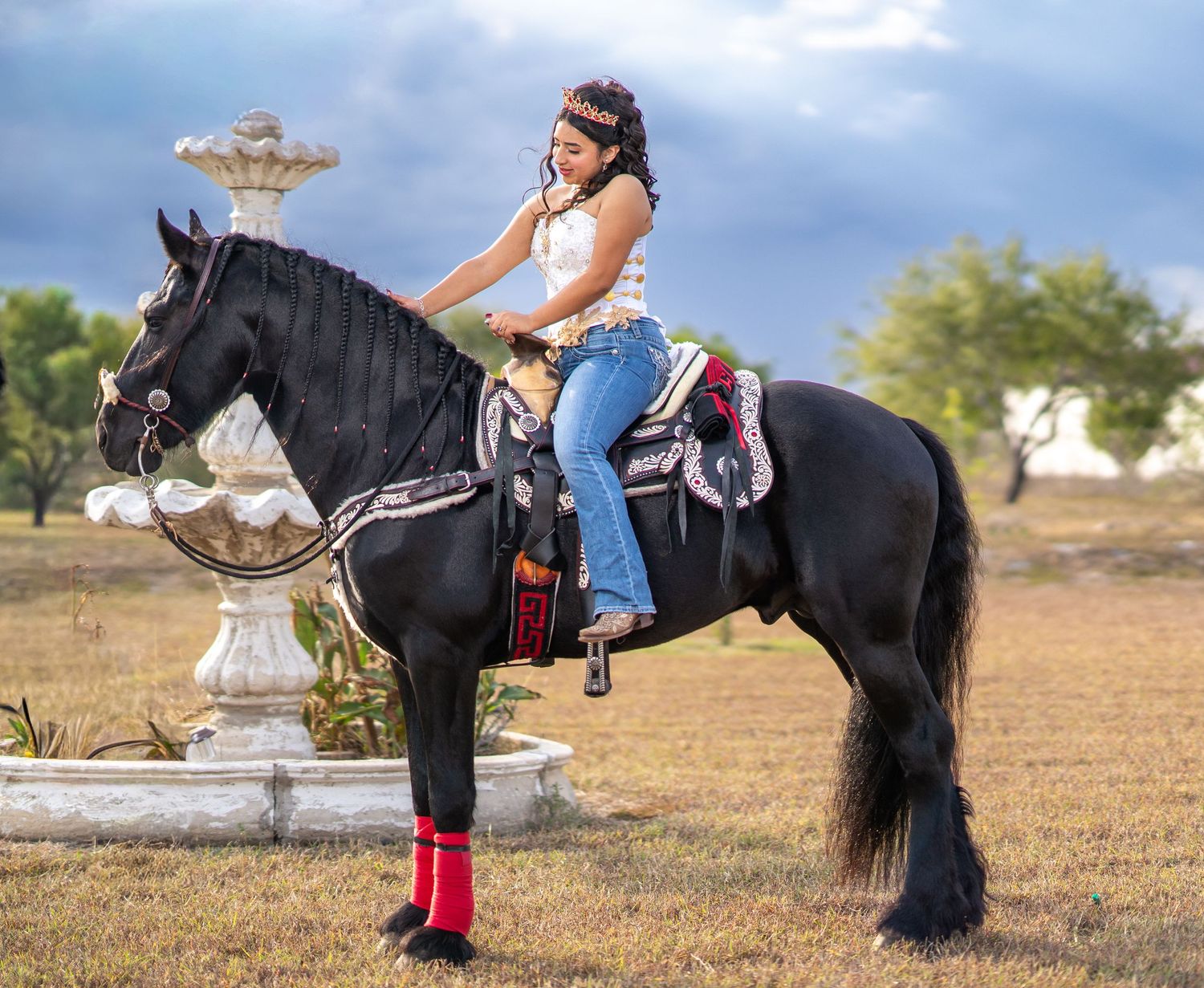 A black horse and someone in western wear pose near a decorative stone fountain in a field.