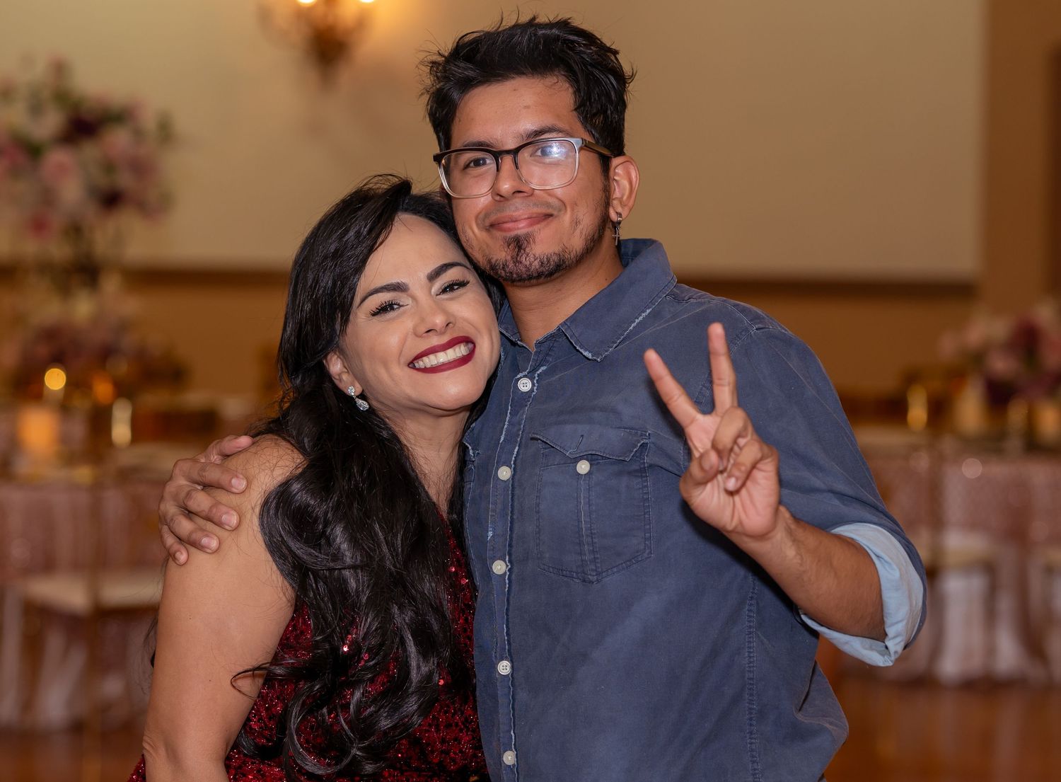 Smiling couple poses together at a quinceañera party, man making a peace sign in a casual blue shirt.