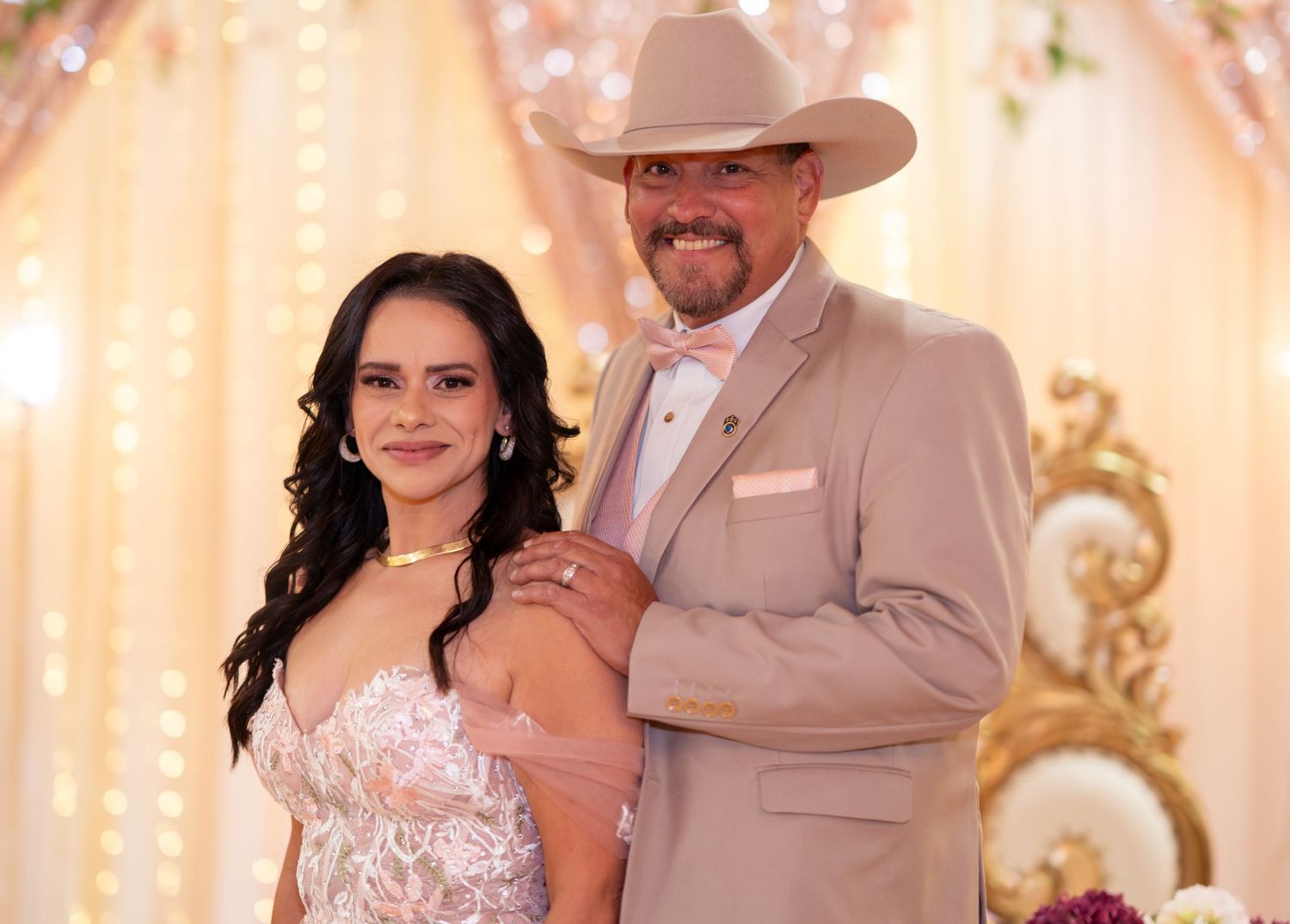 Couple posing together at a quinceañera, man wearing tan suit and cowboy hat, woman in elegant white lace gown.