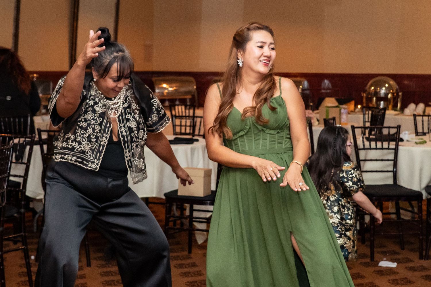Two women dancing joyfully at a formal event, one in a green gown, in an elegantly decorated banquet hall.