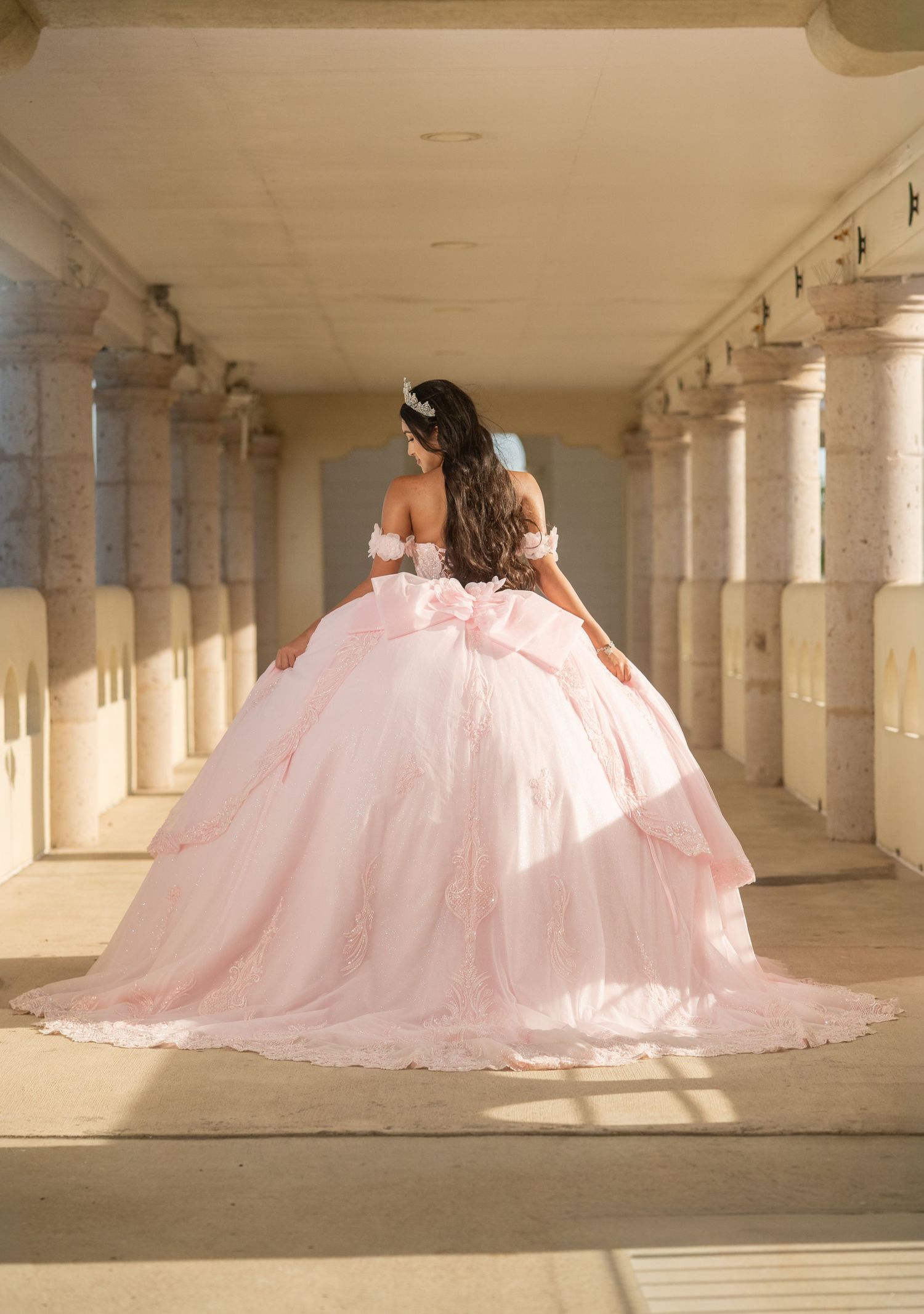 Young woman in a pink ball gown with lace details and bow, posing in a columned hallway for quinceañera.