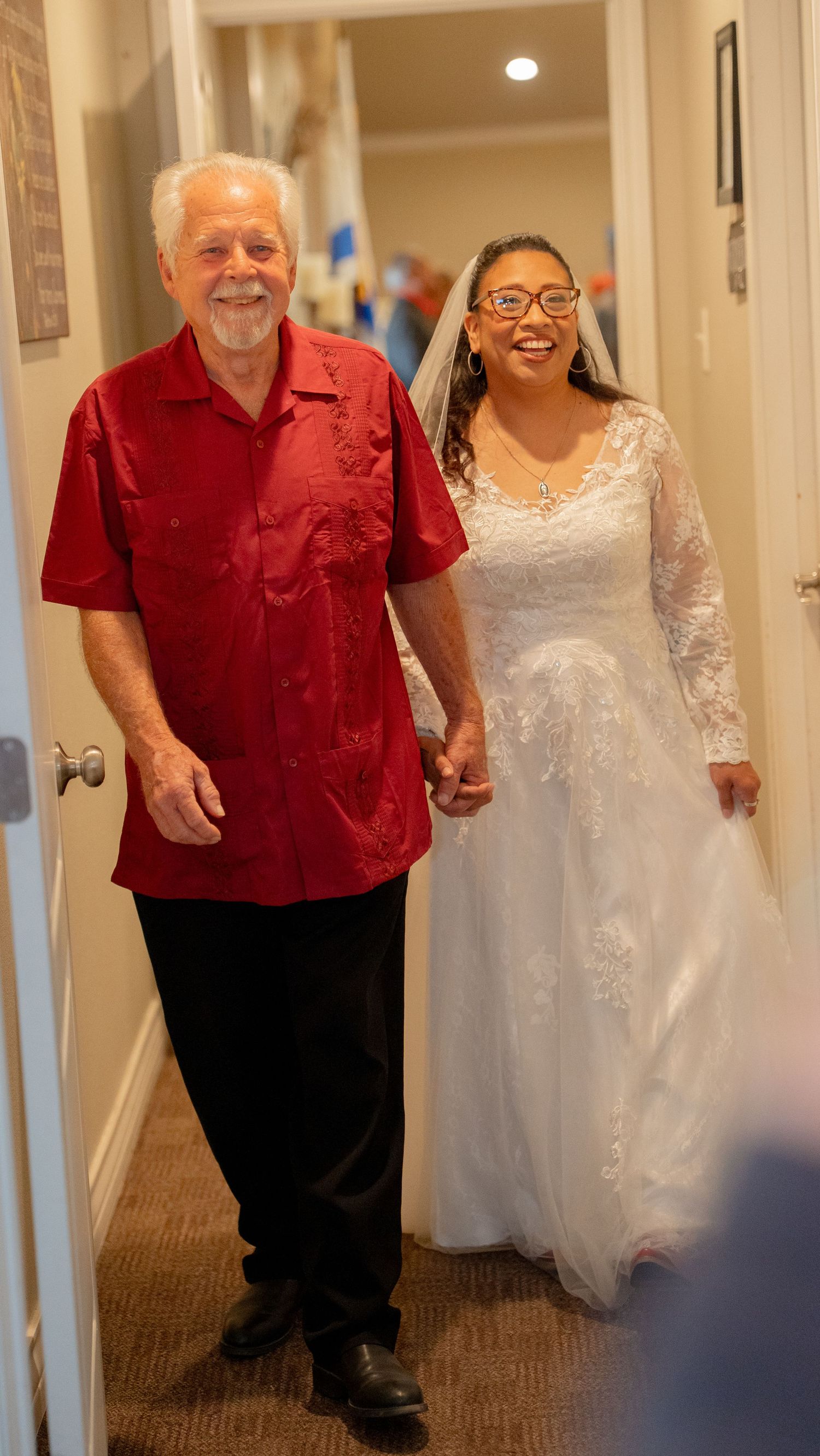 Smiling bride in white lace gown and veil walking hand-in-hand with groom in red shirt down hallway.