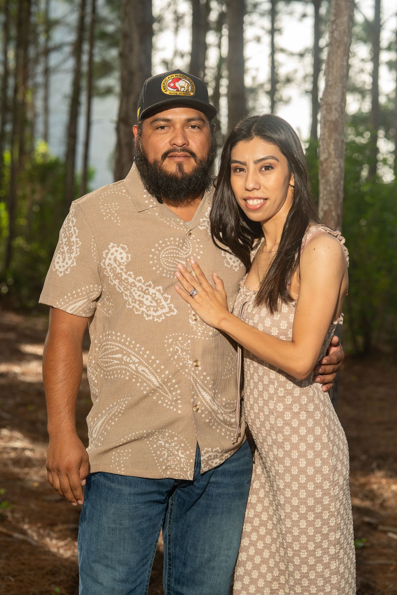 Couple poses together in forest, man in paisley shirt and cap with woman in matching dress, engagement ring visible.