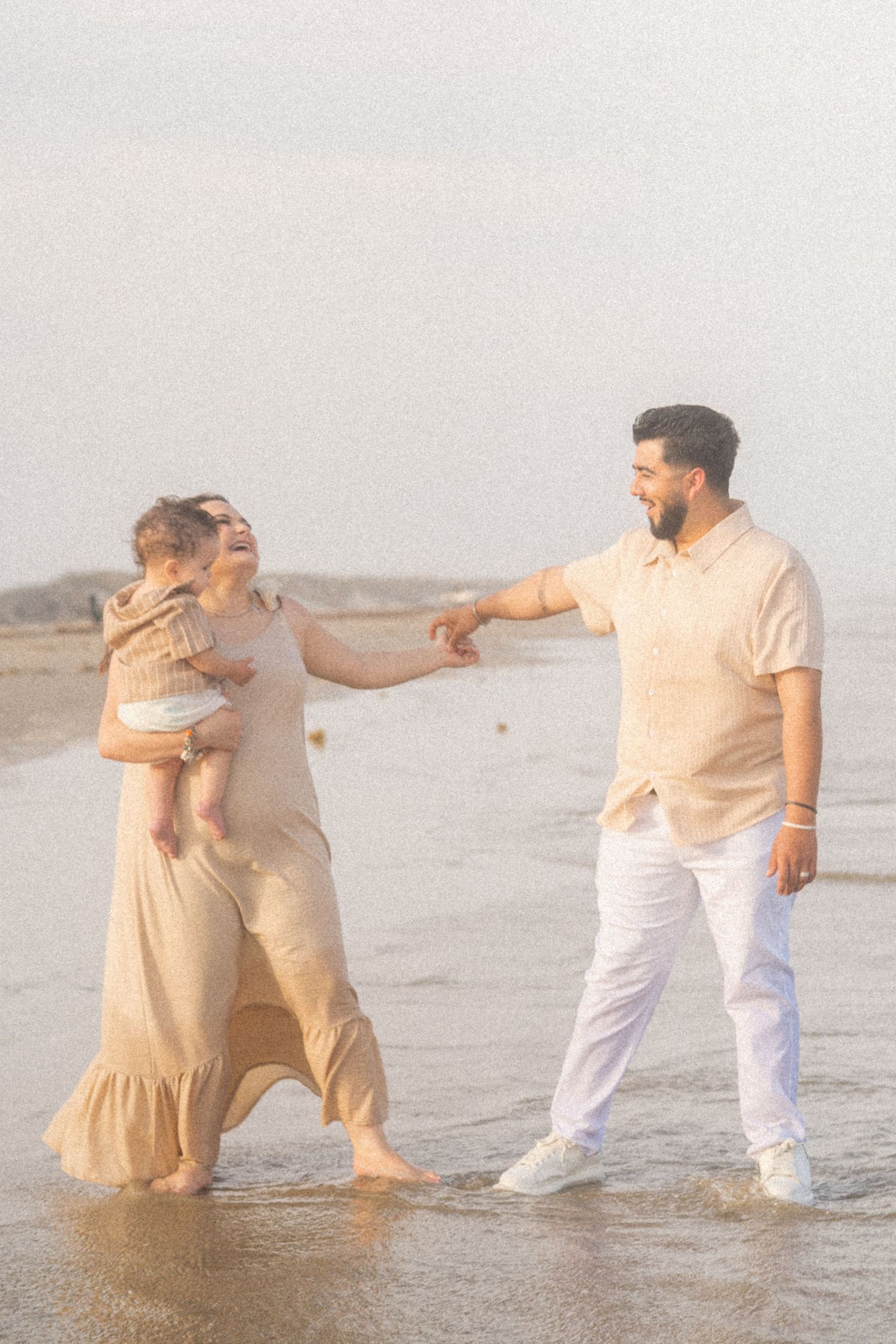 Young family playing in beach waves, woman in maxi dress holding baby while man reaches for her hand.
