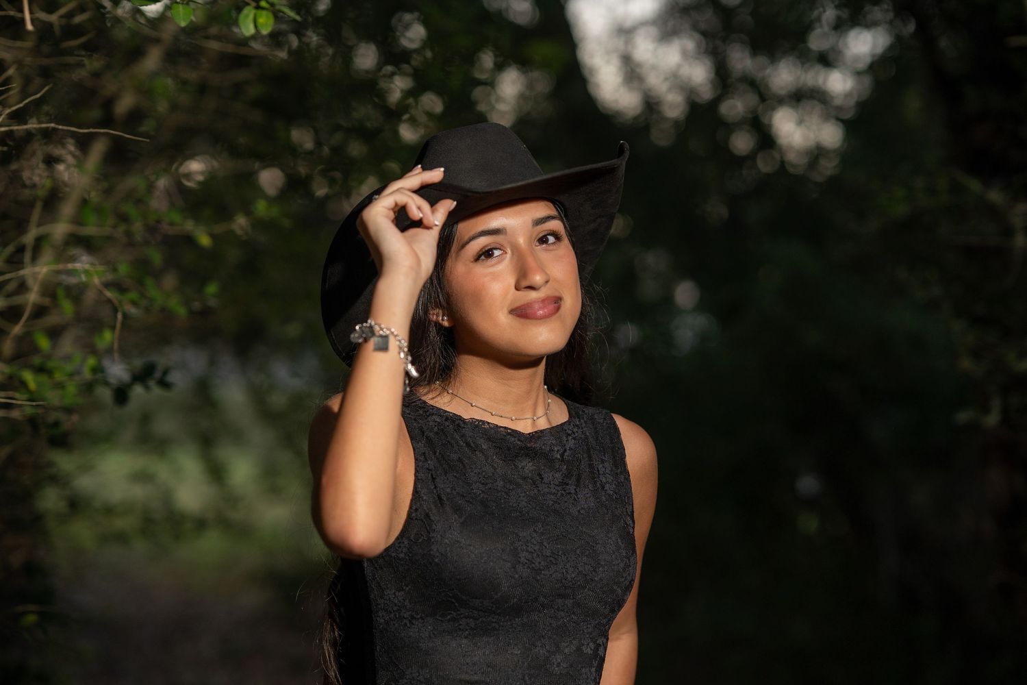 Young Latina teen in black lace dress adjusts black cowboy hat outdoors for quinceañera photos.