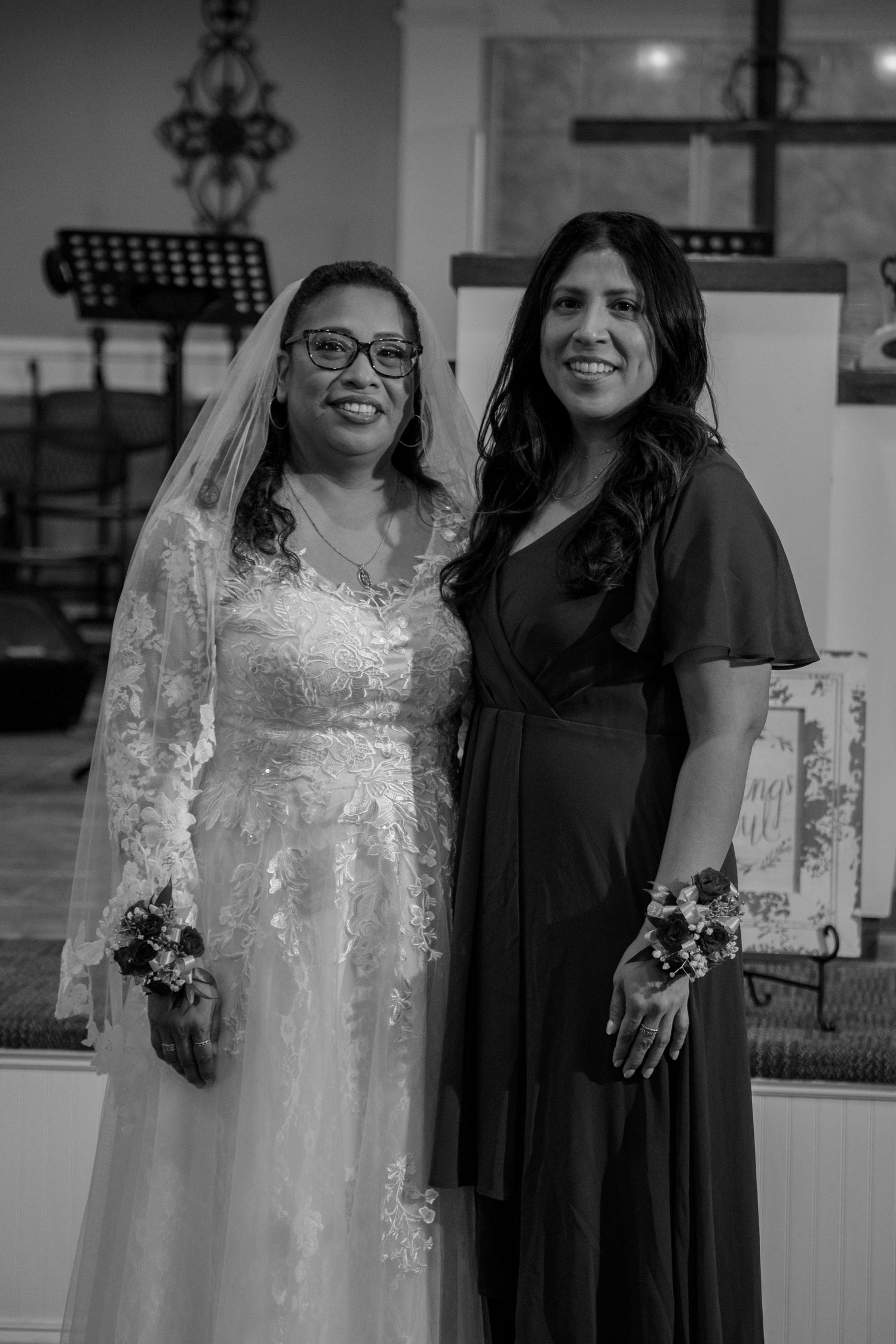 Black and white photo of bride in lace gown and veil posing with female guest in black dress at wedding.