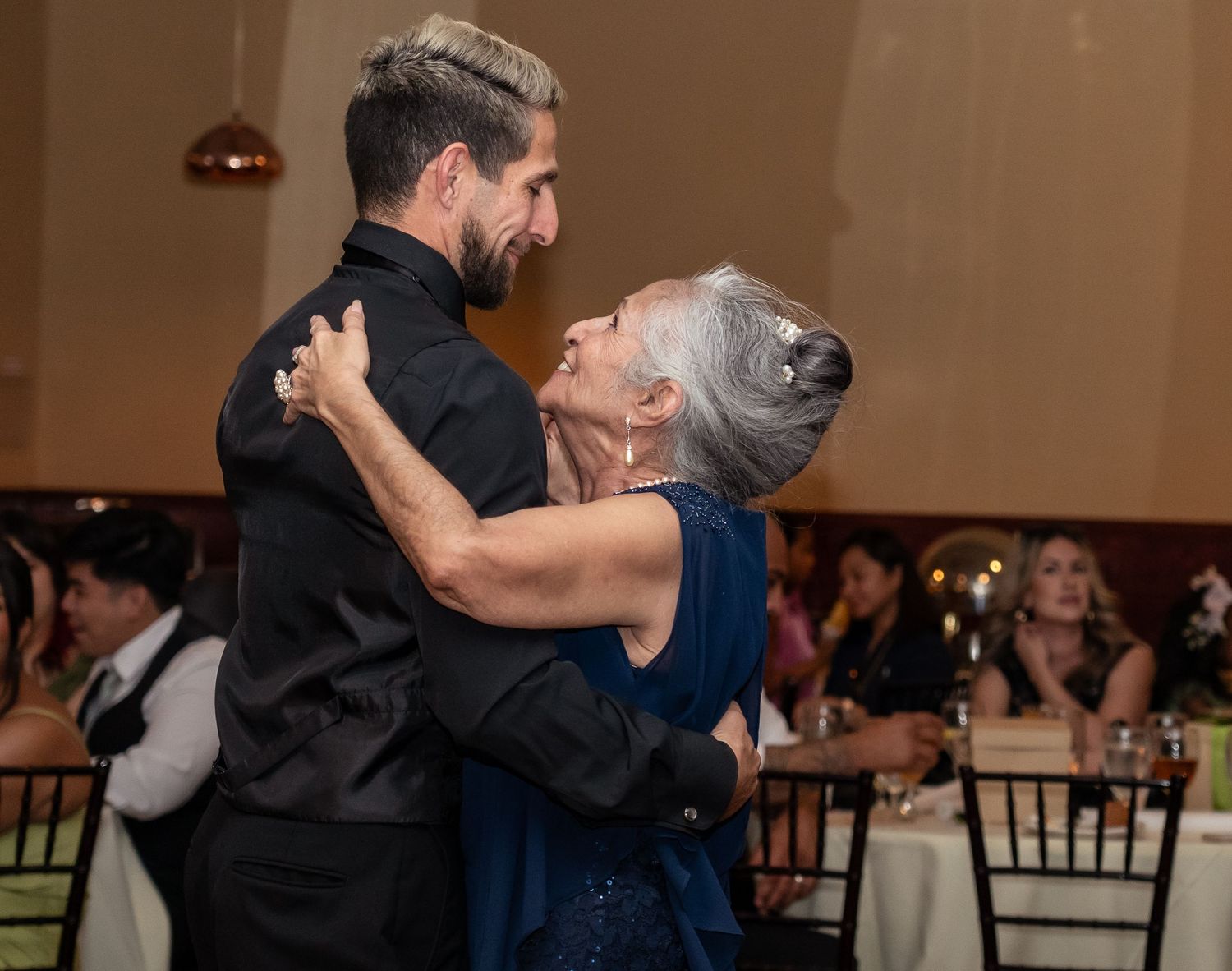 A young man in black dances closely with an older woman in a blue dress at an elegant event.