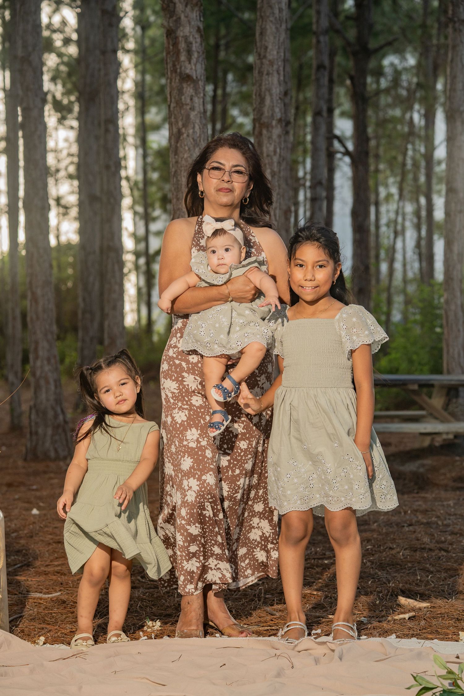 A mother holds her baby while standing with two young girls in green dresses among tall pine trees outdoors.