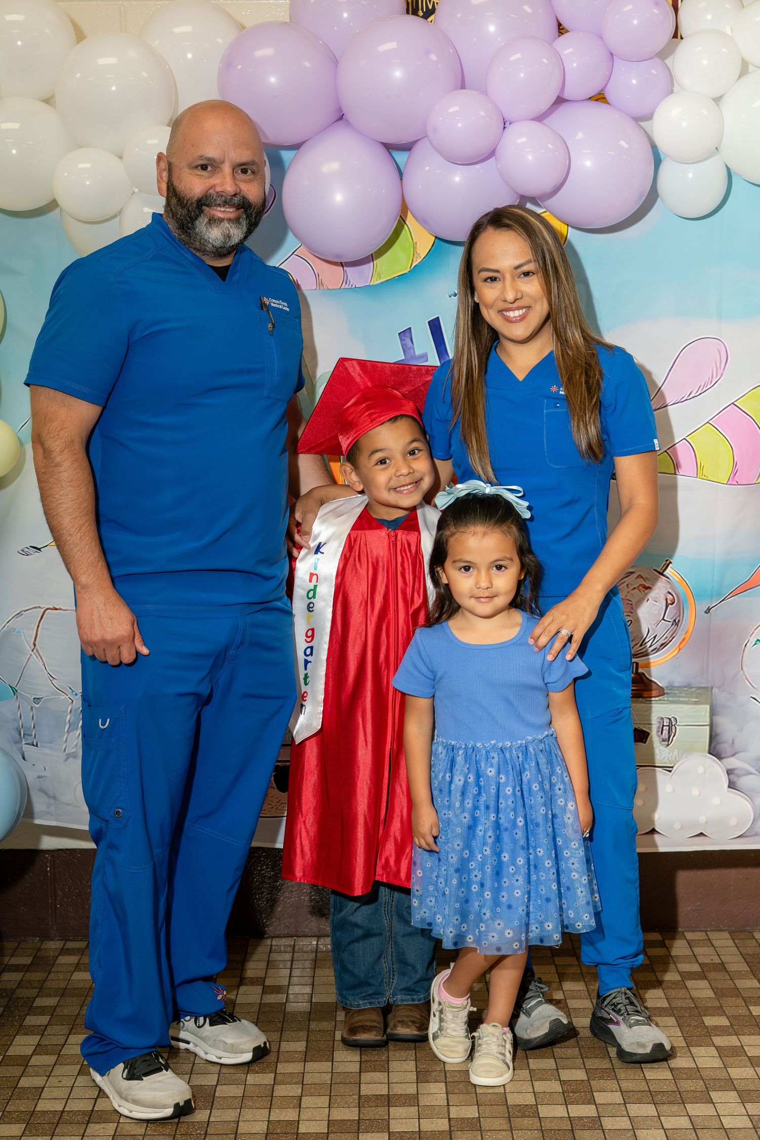 Family of four poses at graduation celebration, child in red cap and gown with balloon backdrop.